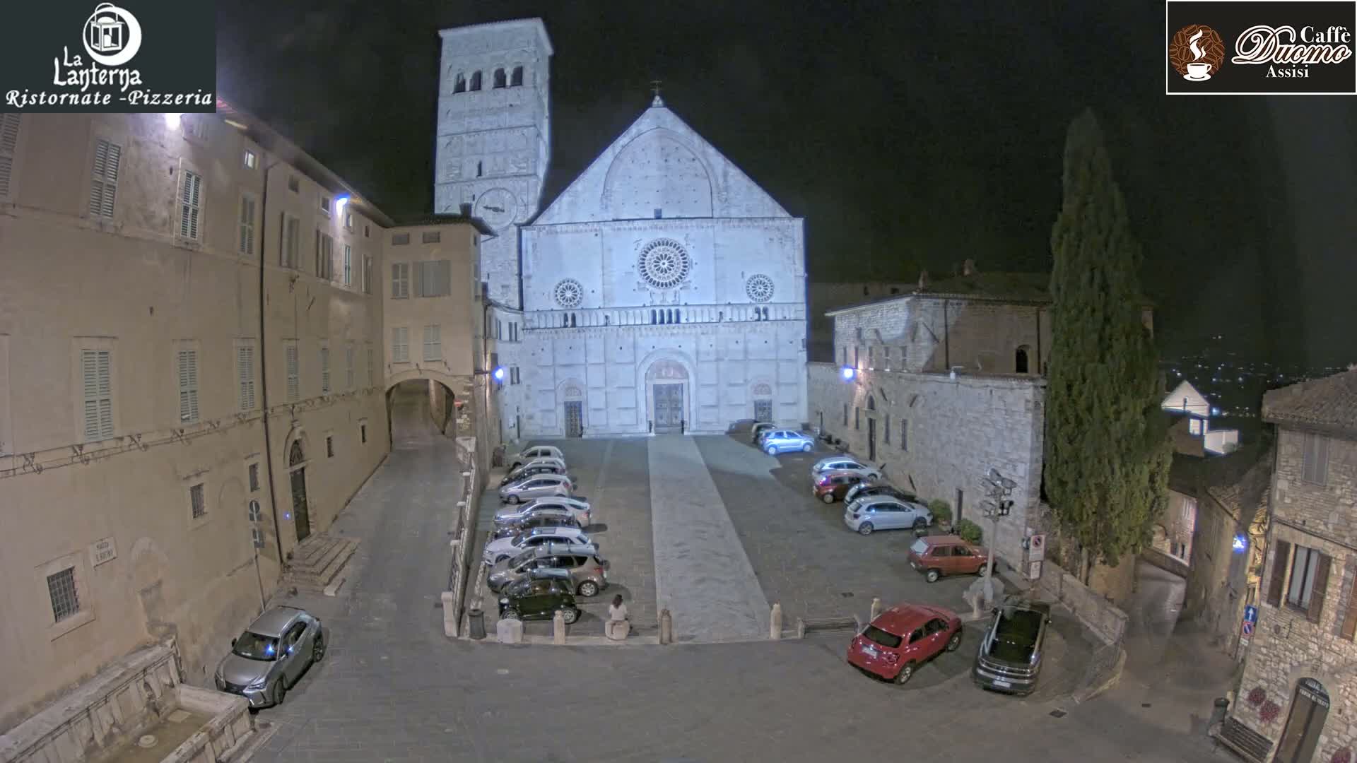 At night, a large stone church is situated in a town square with cars parked in front of it, under a clear sky.