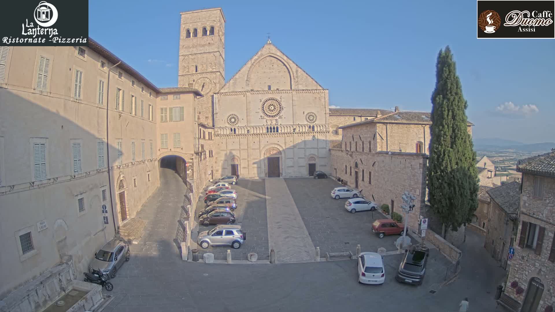 A large stone cathedral sits in a town square with several cars parked, under a clear, sunny sky.