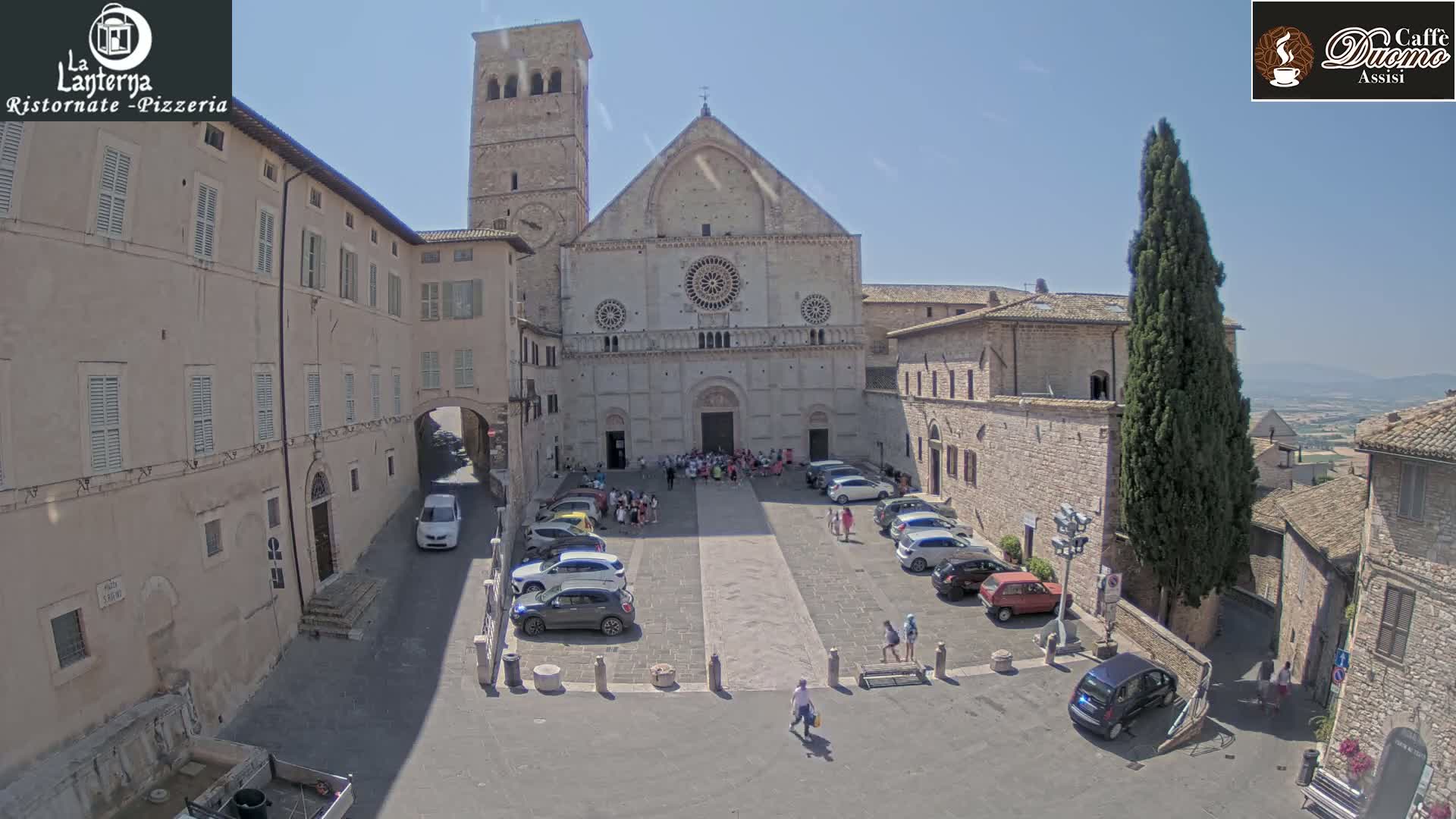 A large stone cathedral is seen in a town square on a sunny day, surrounded by parked cars and several people.