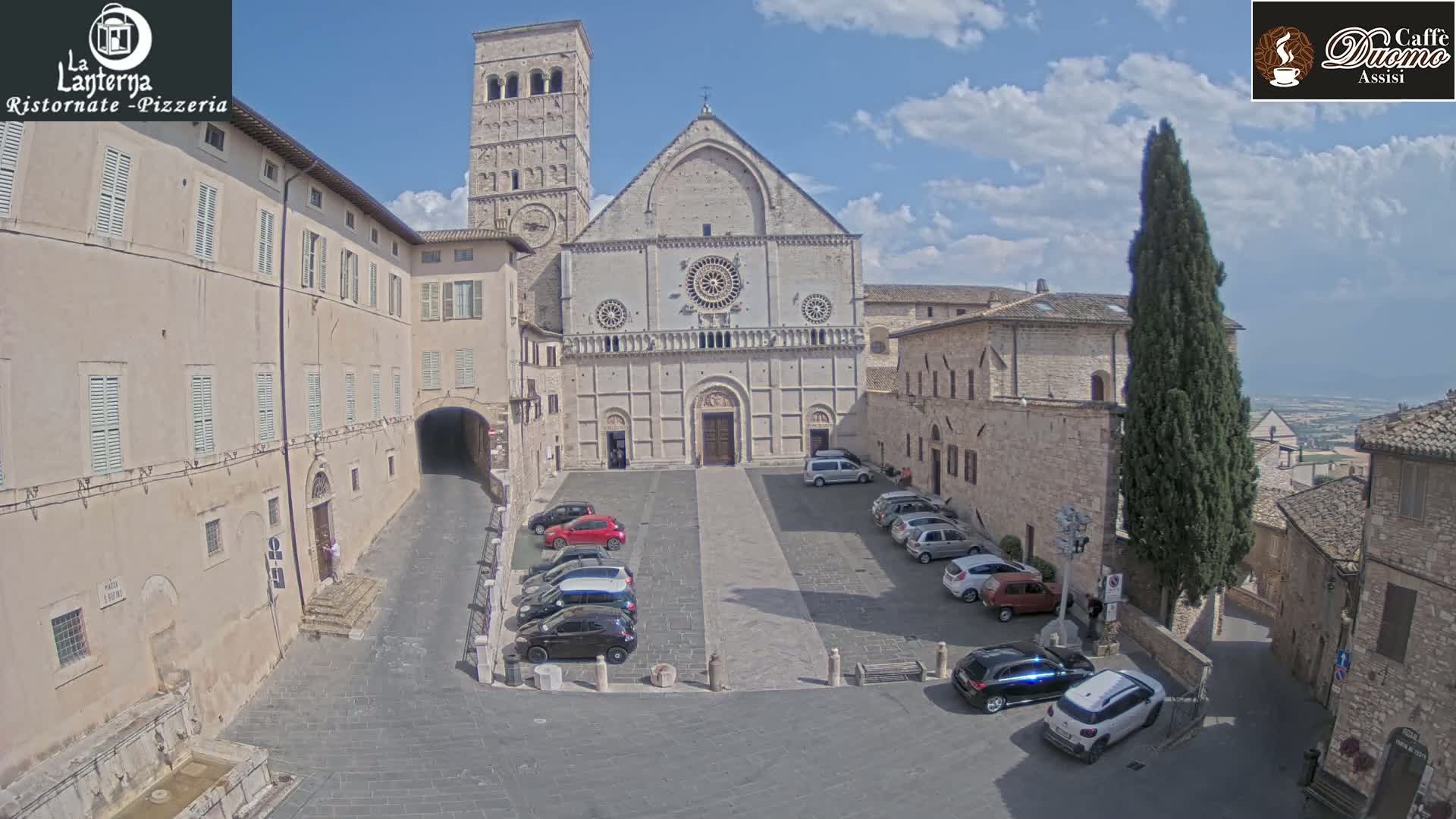 A sunny day in a stone plaza with parked cars in front of a large cathedral and adjacent buildings.