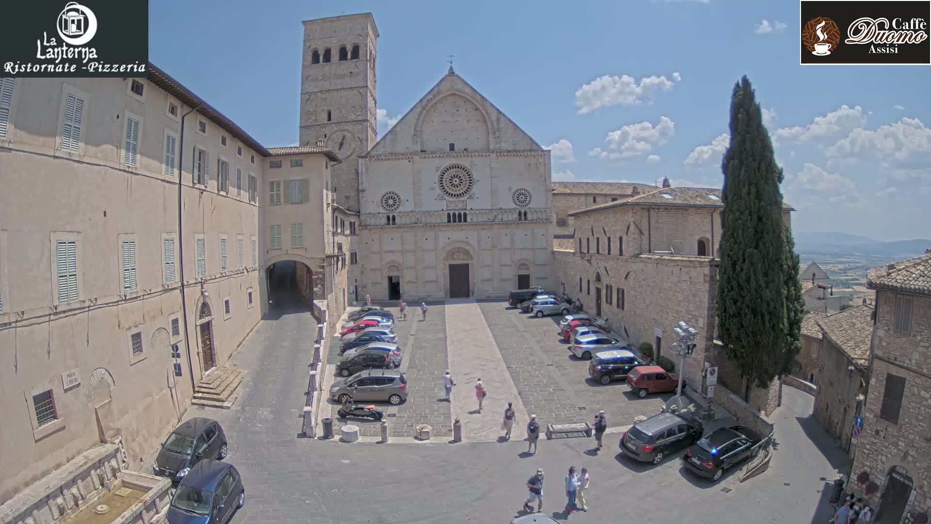 A sunny day in a European town square shows a large stone church with cars parked in front, surrounded by buildings, and a few people walking around.