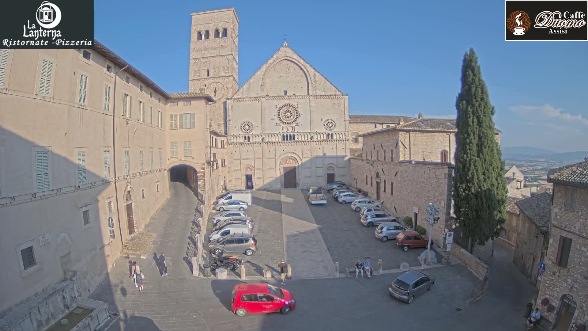 A large stone cathedral sits in a town square with cars parked around it, under a clear, sunny sky.