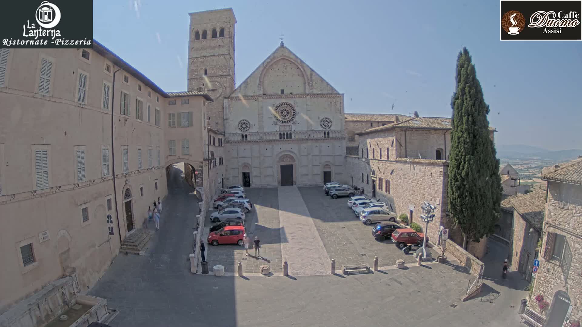 A large stone church faces a paved square filled with parked cars under a sunny sky, surrounded by stone buildings.