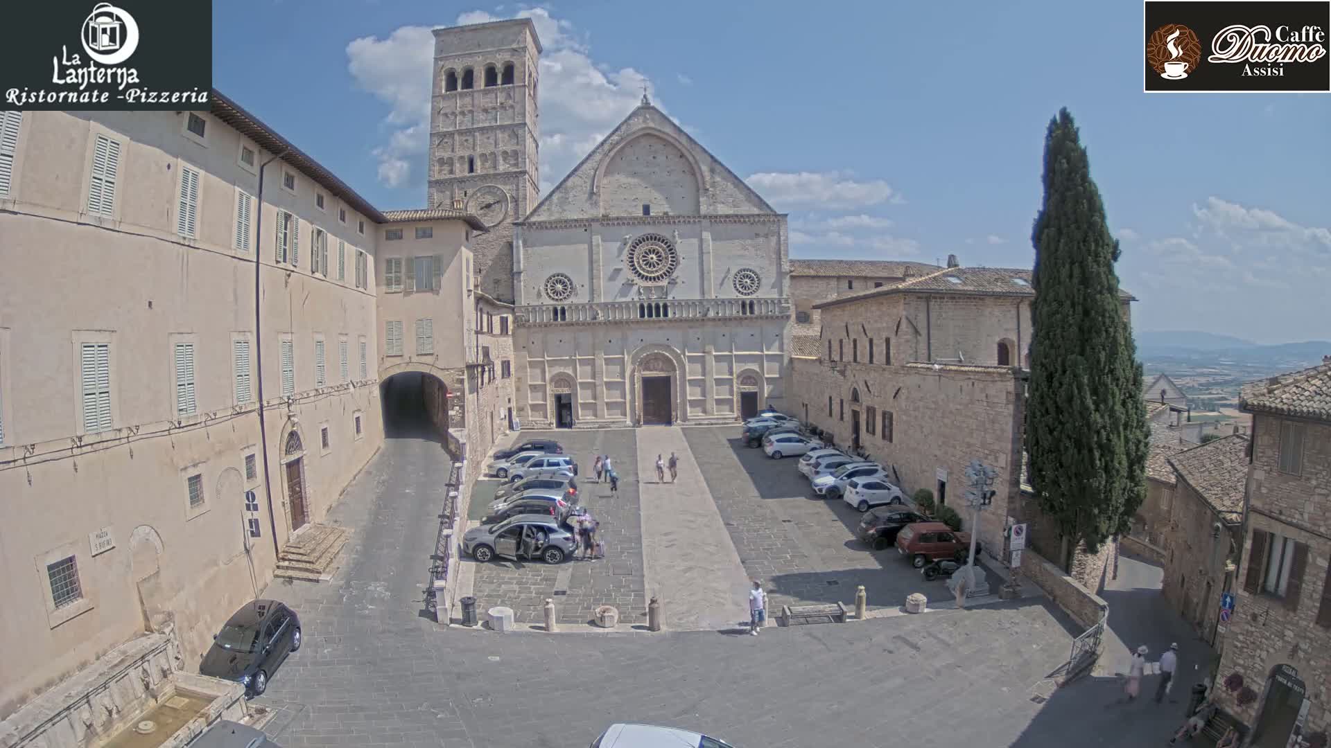 A sunny day reveals a town square with parked cars in front of a large stone cathedral and adjacent buildings.
