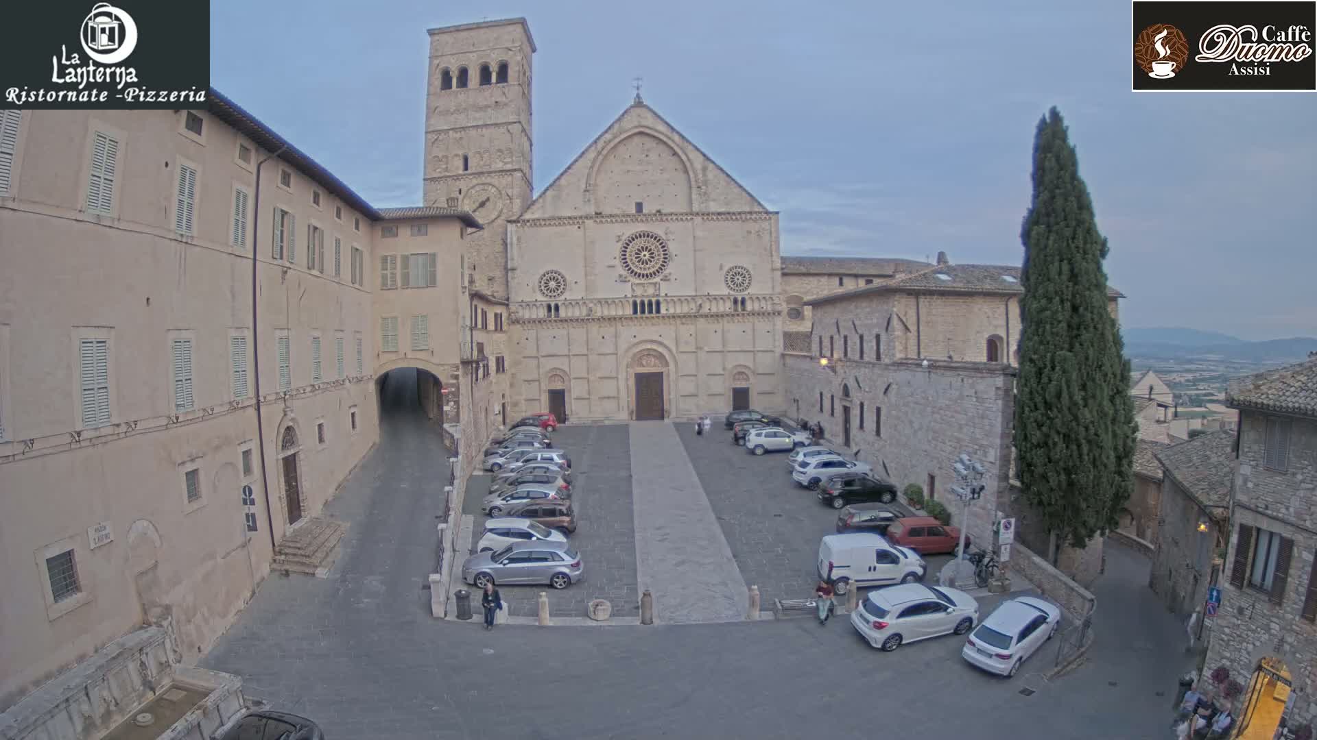 A large stone cathedral stands in a plaza with parked cars under a cloudy sky.