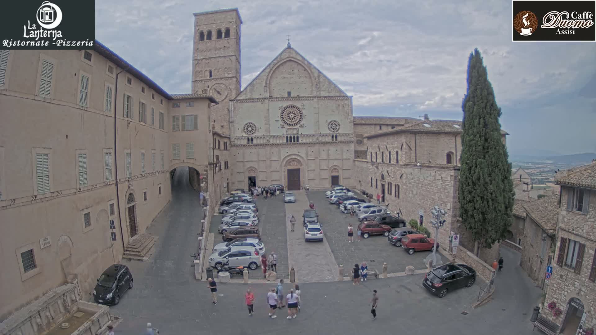 A large stone cathedral is situated in a town square with cars parked around it, under an overcast sky.