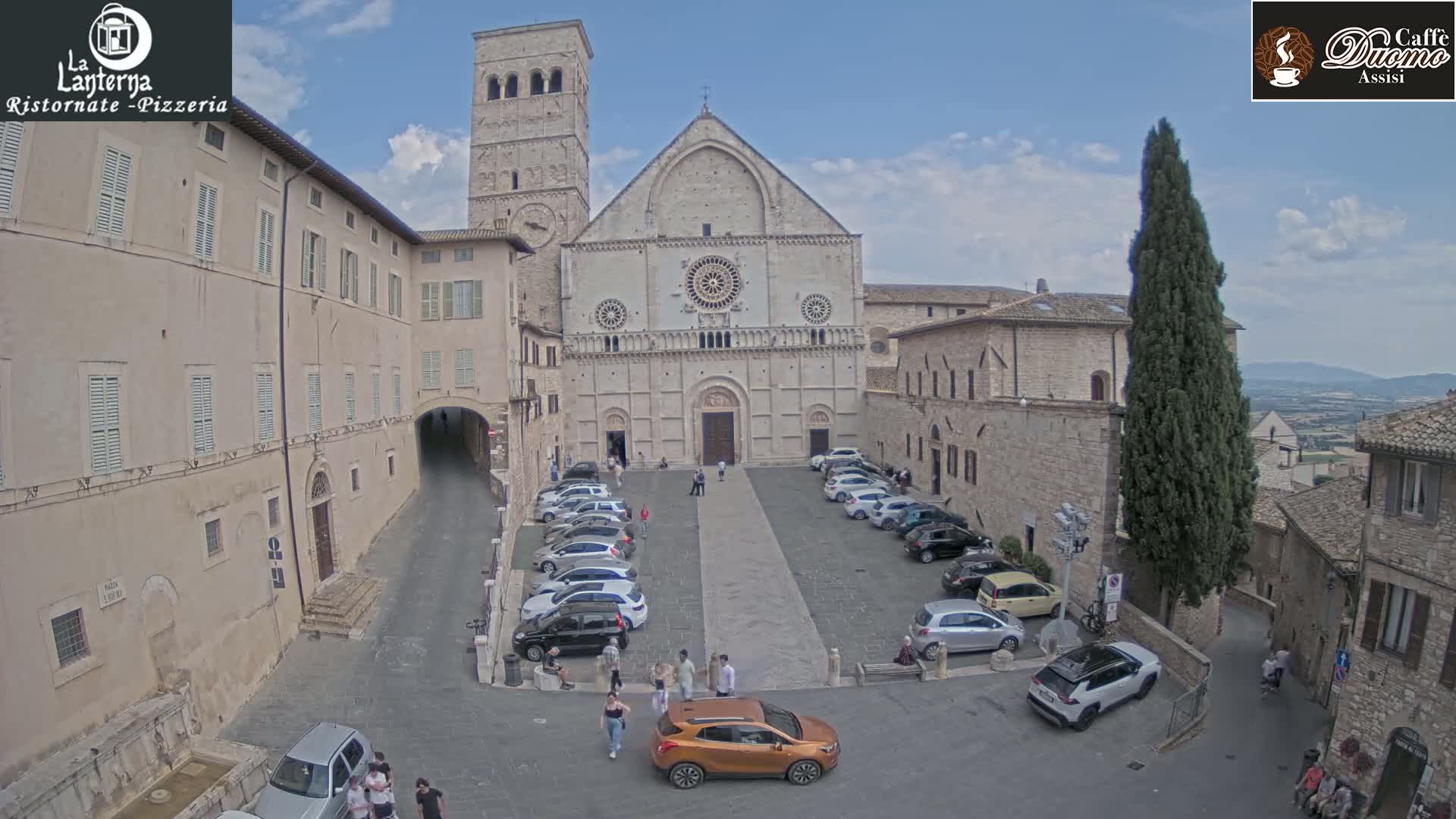 A large stone cathedral sits in a town square, surrounded by buildings and parked cars under a partly cloudy sky.