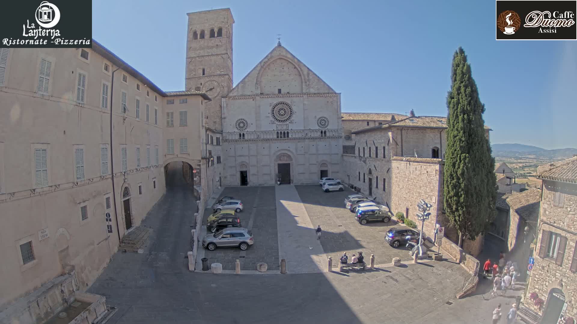 A large stone church and adjacent buildings are situated in a town square on a sunny day, with several cars parked in the square and people walking around.