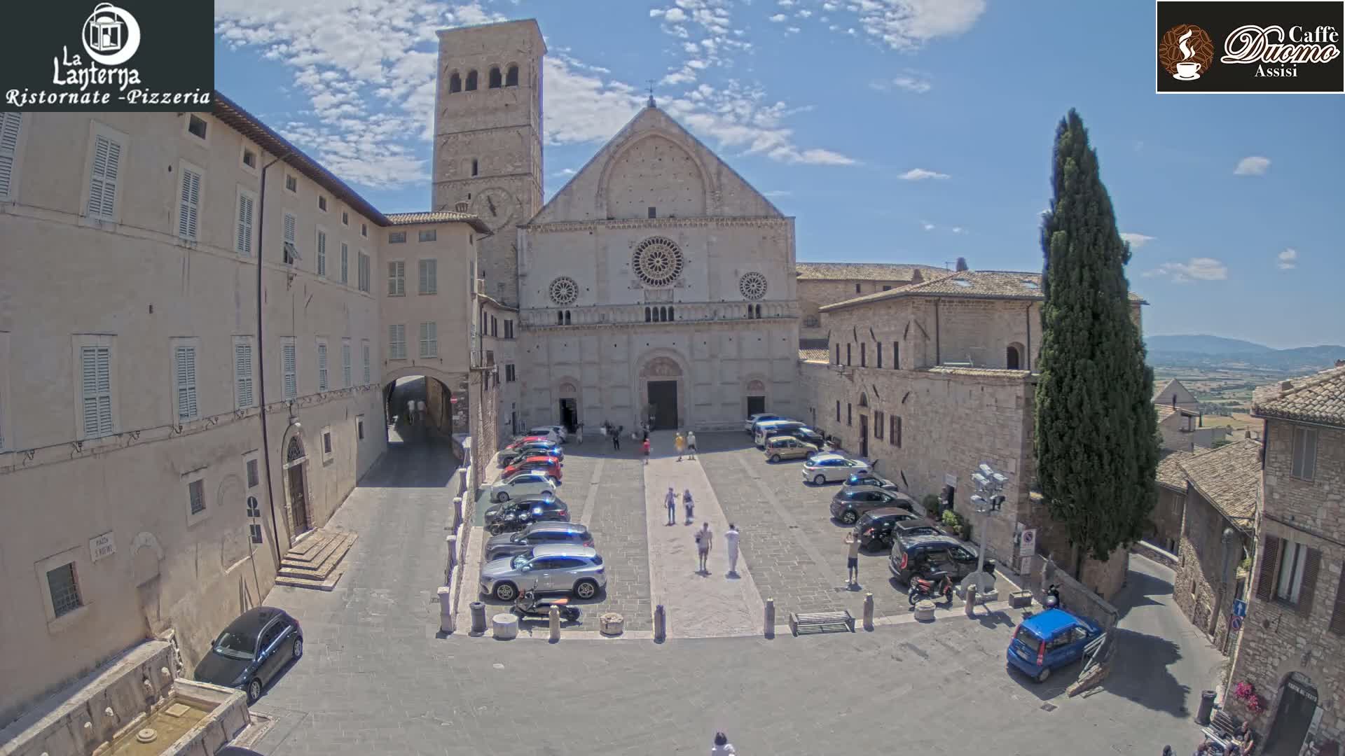 A large stone church and adjacent buildings are situated in a town square on a sunny day, with several cars parked in the square and people walking around.