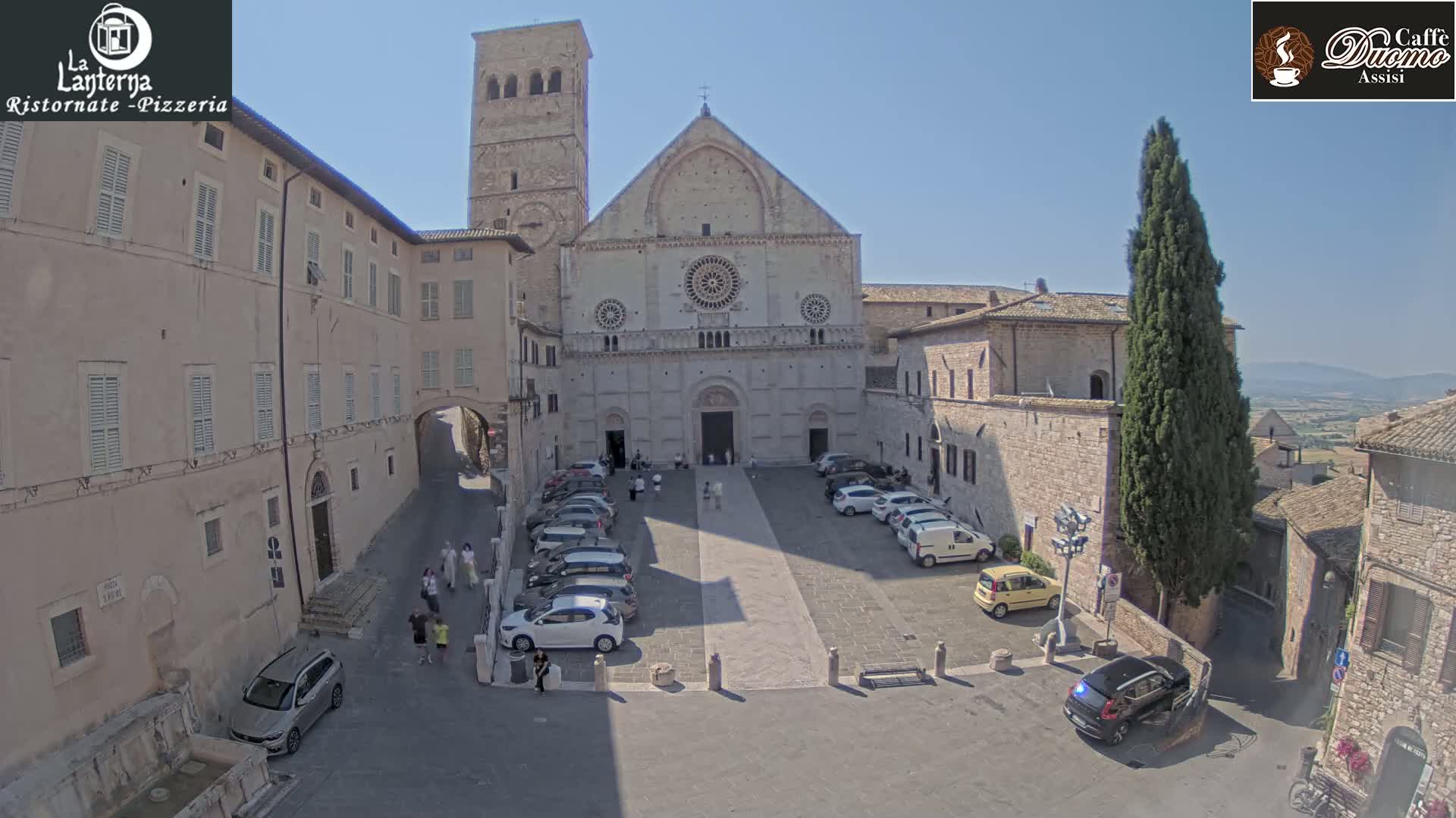 A large stone cathedral is situated in a town square with cars parked around it, under sunny skies.