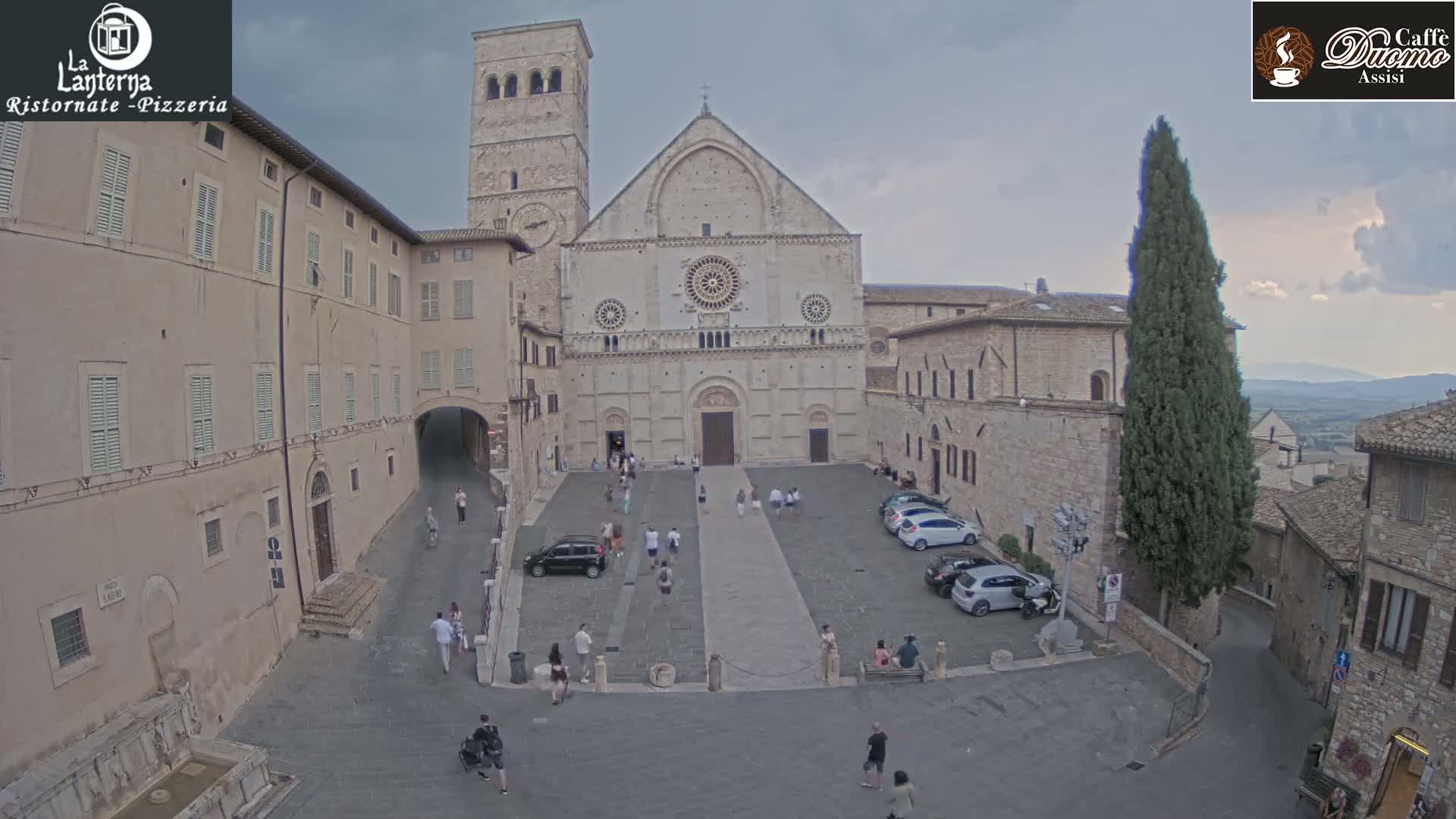 A large stone cathedral is situated in a town square with cars parked around it, under sunny skies.