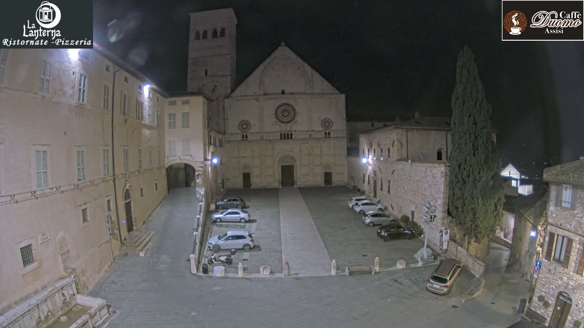 On a clear night, a historic Italian piazza is illuminated, centered by a grand stone basilica with a large rose window and bell tower, surrounded by traditional buildings, parked cars, and an old fountain.