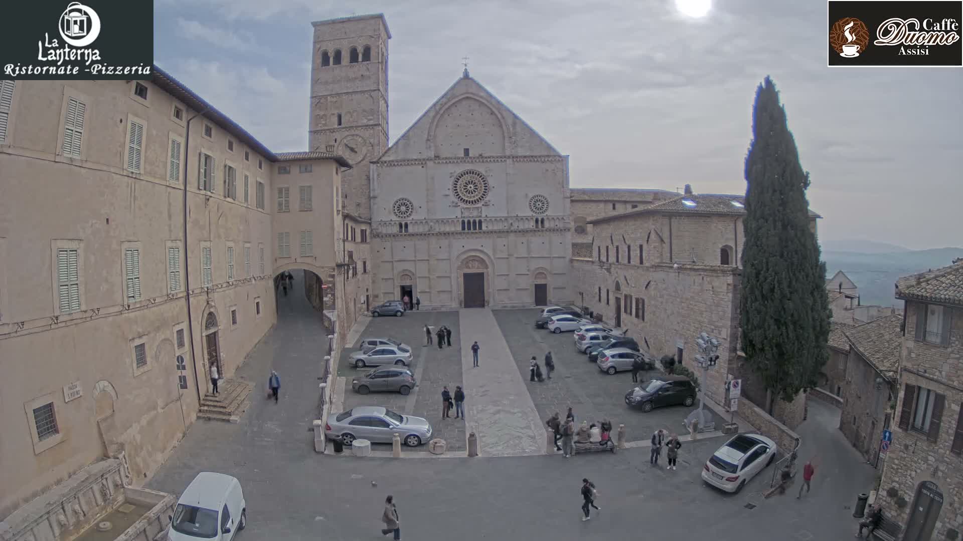 On an overcast day, a wide view captures a historic Italian town square featuring a prominent stone cathedral with a tall bell tower, surrounded by old buildings, parked cars, and pedestrians milling about.