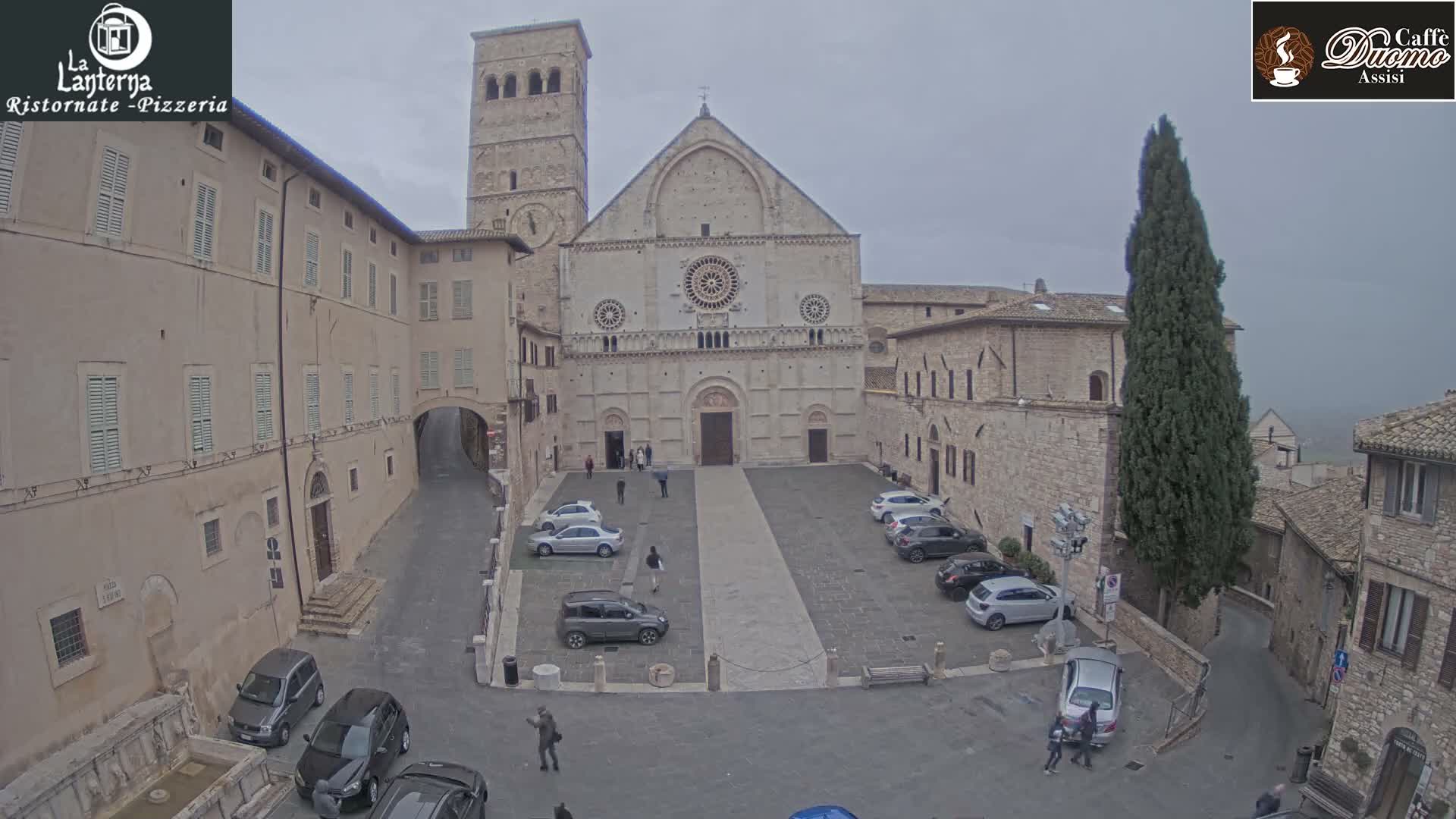 On an overcast day, a wide view captures a historic Italian town square featuring a prominent stone cathedral with a tall bell tower, surrounded by old buildings, parked cars, and pedestrians milling about.