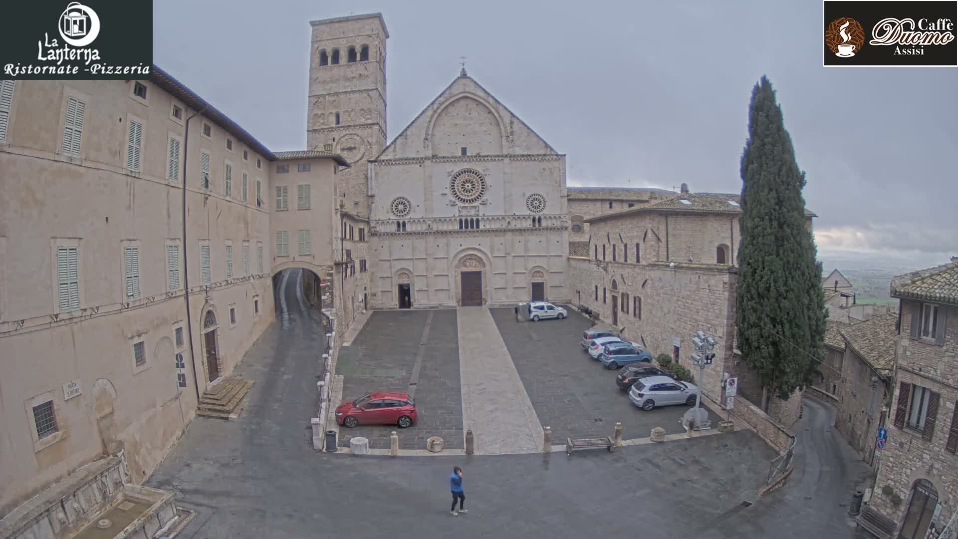 An overcast sky hangs over a grand stone basilica with a tall bell tower, overlooking a paved plaza flanked by historic buildings and a cypress tree, with a few cars parked and a solitary figure walking.