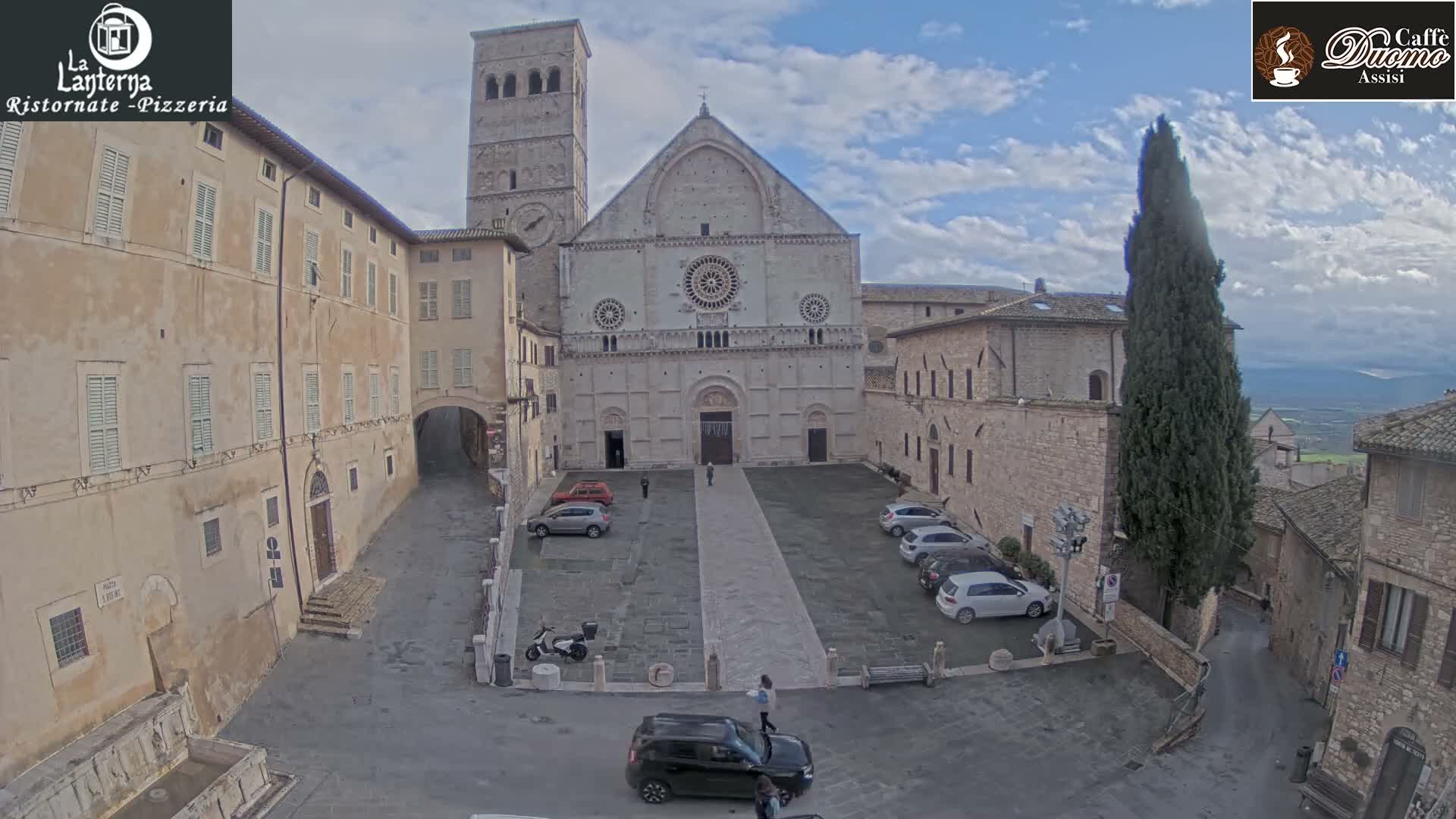 An overcast sky hangs over a grand stone basilica with a tall bell tower, overlooking a paved plaza flanked by historic buildings and a cypress tree, with a few cars parked and a solitary figure walking.
