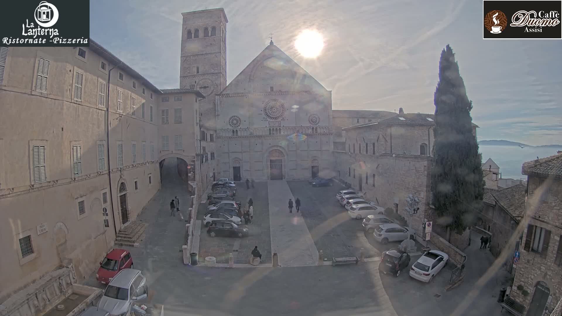 A bright, sunny day illuminates a European town square with a large stone basilica and bell tower at its center, surrounded by historic buildings, people, and parked cars, with a hazy mountain range in the distant background.