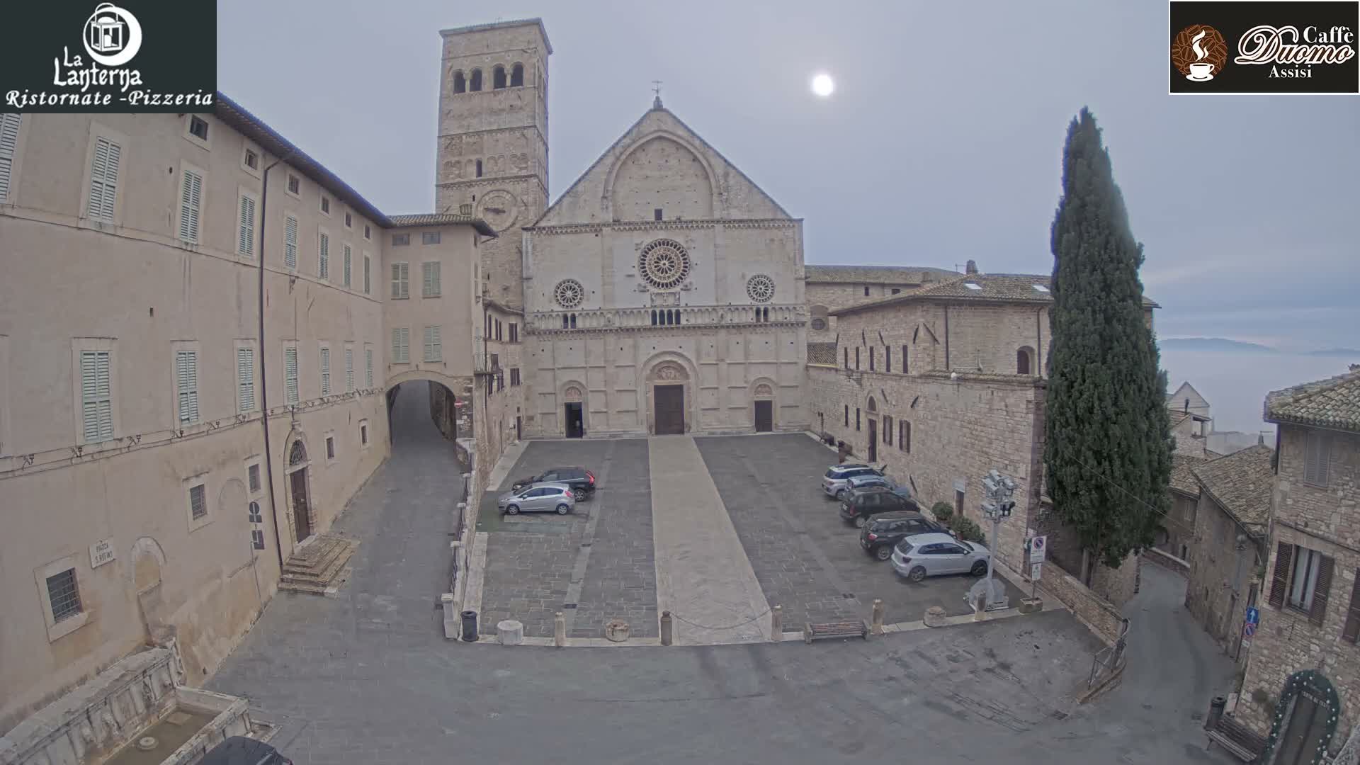 A historic stone basilica with a large bell tower overlooks a cobblestone piazza featuring parked cars and surrounding old buildings, all under an overcast and hazy sky with the sun appearing as a diffused disc.