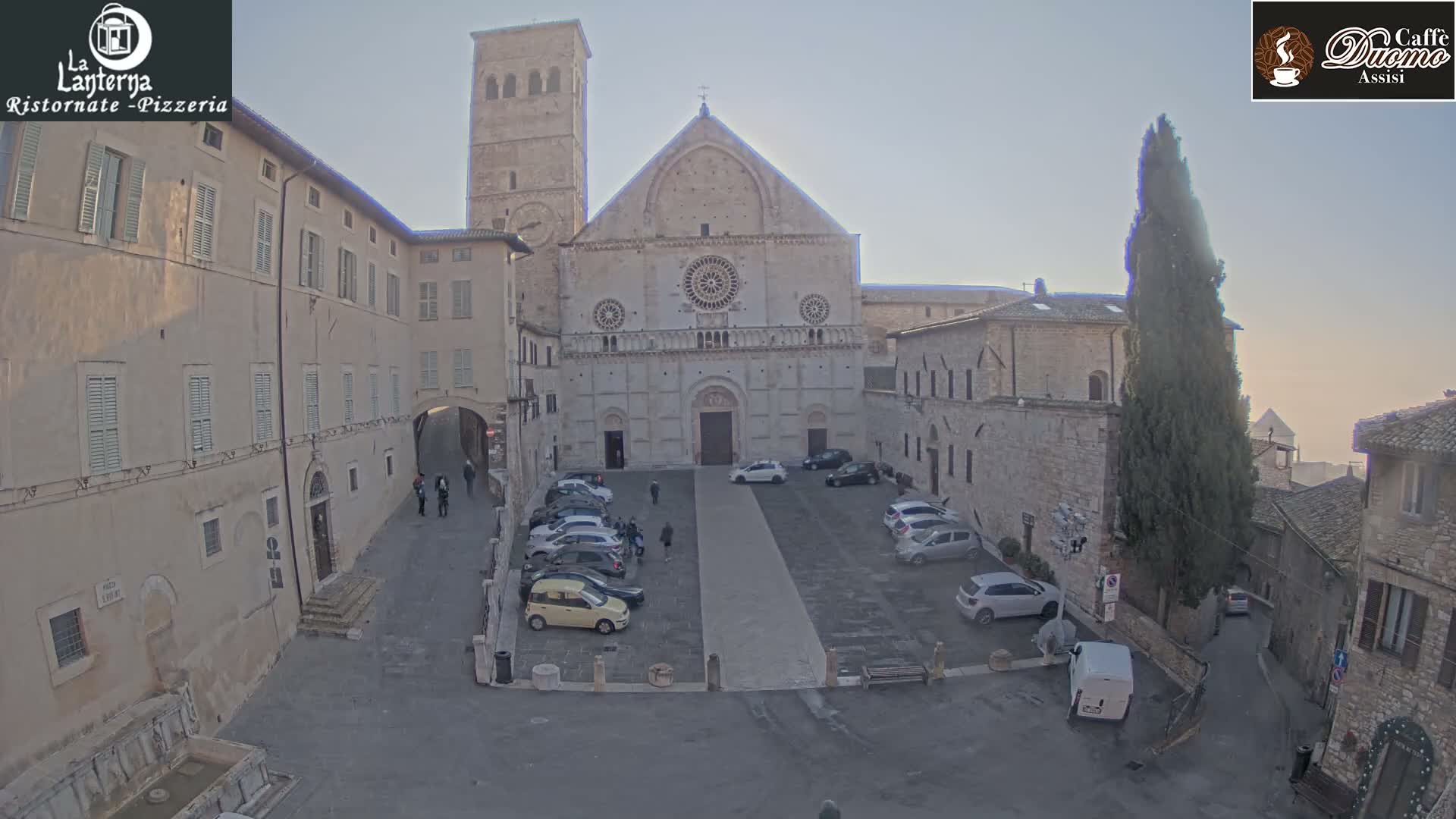 A historic stone basilica with a large bell tower overlooks a cobblestone piazza featuring parked cars and surrounding old buildings, all under an overcast and hazy sky with the sun appearing as a diffused disc.