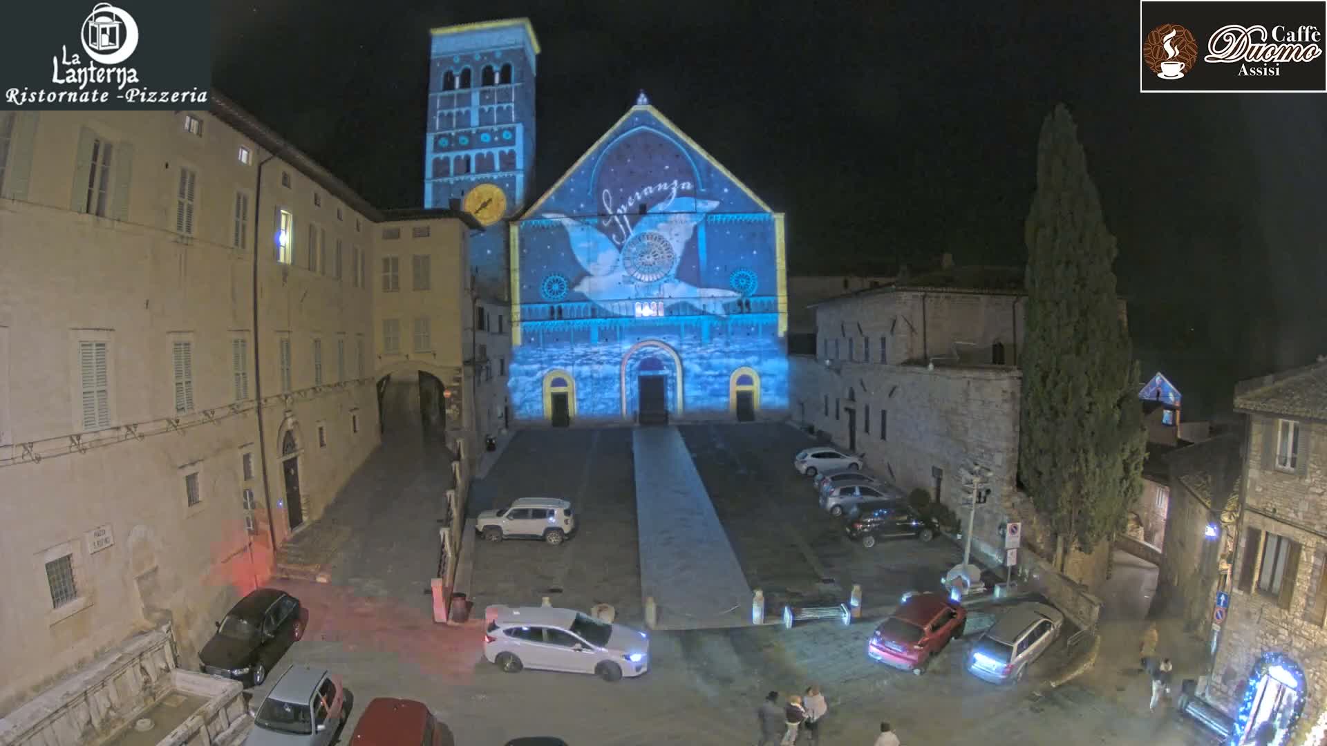 A historic stone basilica with a large bell tower overlooks a cobblestone piazza featuring parked cars and surrounding old buildings, all under an overcast and hazy sky with the sun appearing as a diffused disc.