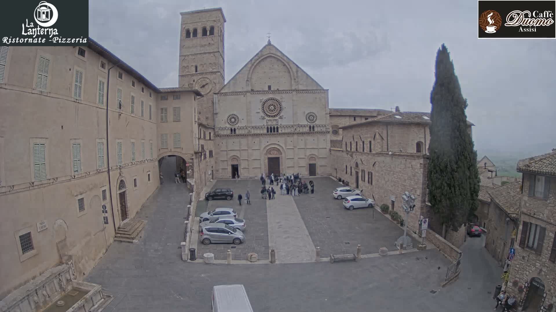 A historic stone basilica with a large bell tower overlooks a cobblestone piazza featuring parked cars and surrounding old buildings, all under an overcast and hazy sky with the sun appearing as a diffused disc.