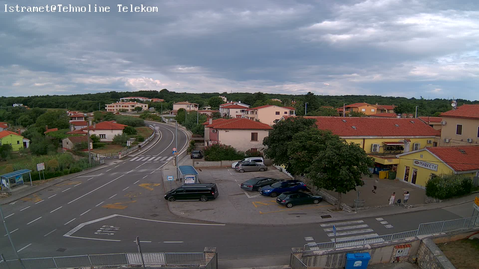 A small town with overcast skies shows several cars parked near a market and a road curving through the residential area.
