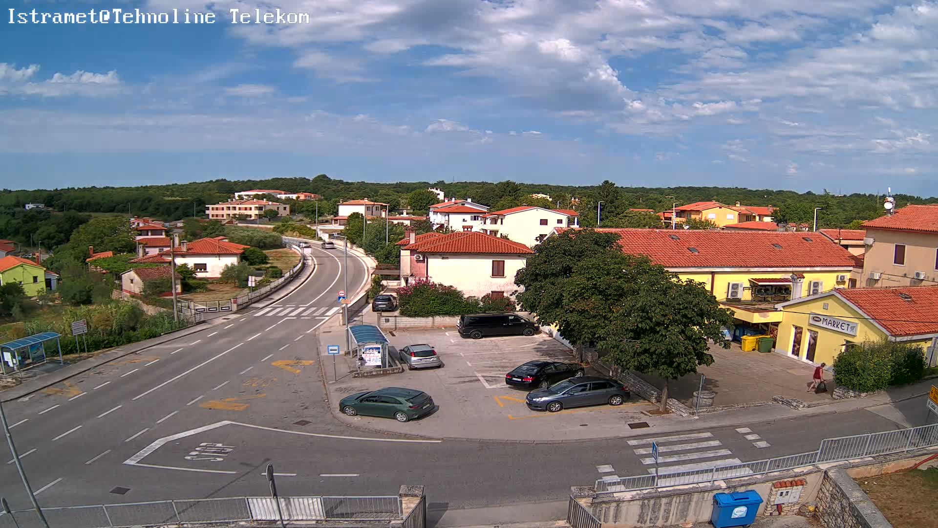 A sunny day shows a small town's street scene with several cars parked near a market and other buildings, and a road curving through the area.
