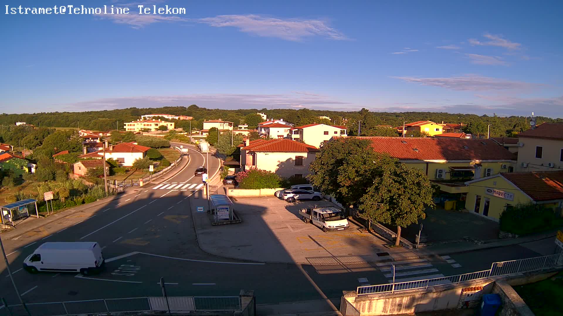 A sunny day shows a small town's street scene with several cars parked near a market and other buildings, and a road curving through the area.