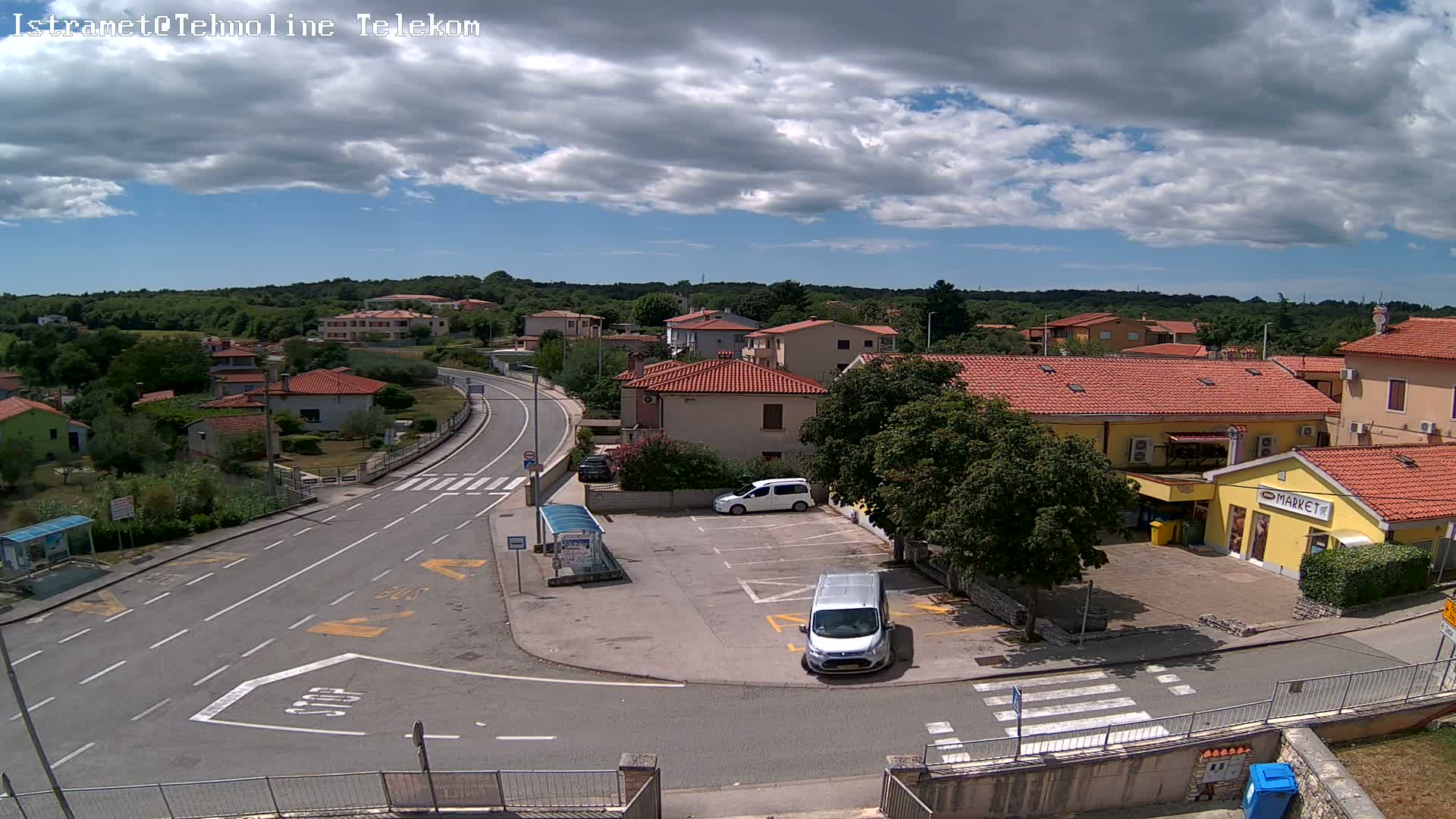 A sunny day shows a small town's street scene with several cars parked near a market and other buildings, and a road curving through the area.