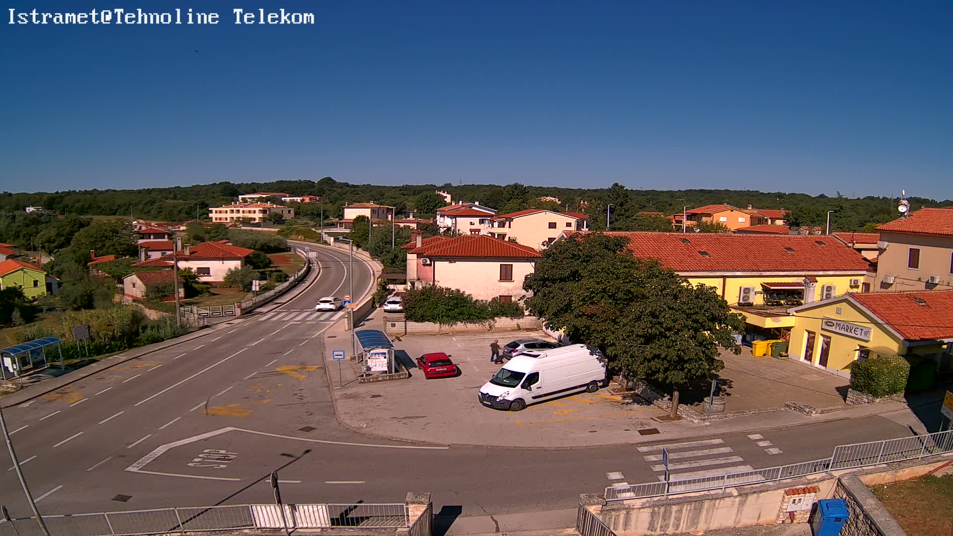 A high-angle view on a sunny day shows a small town with red-tiled roofs, a road curving through it, and several vehicles parked near a building.