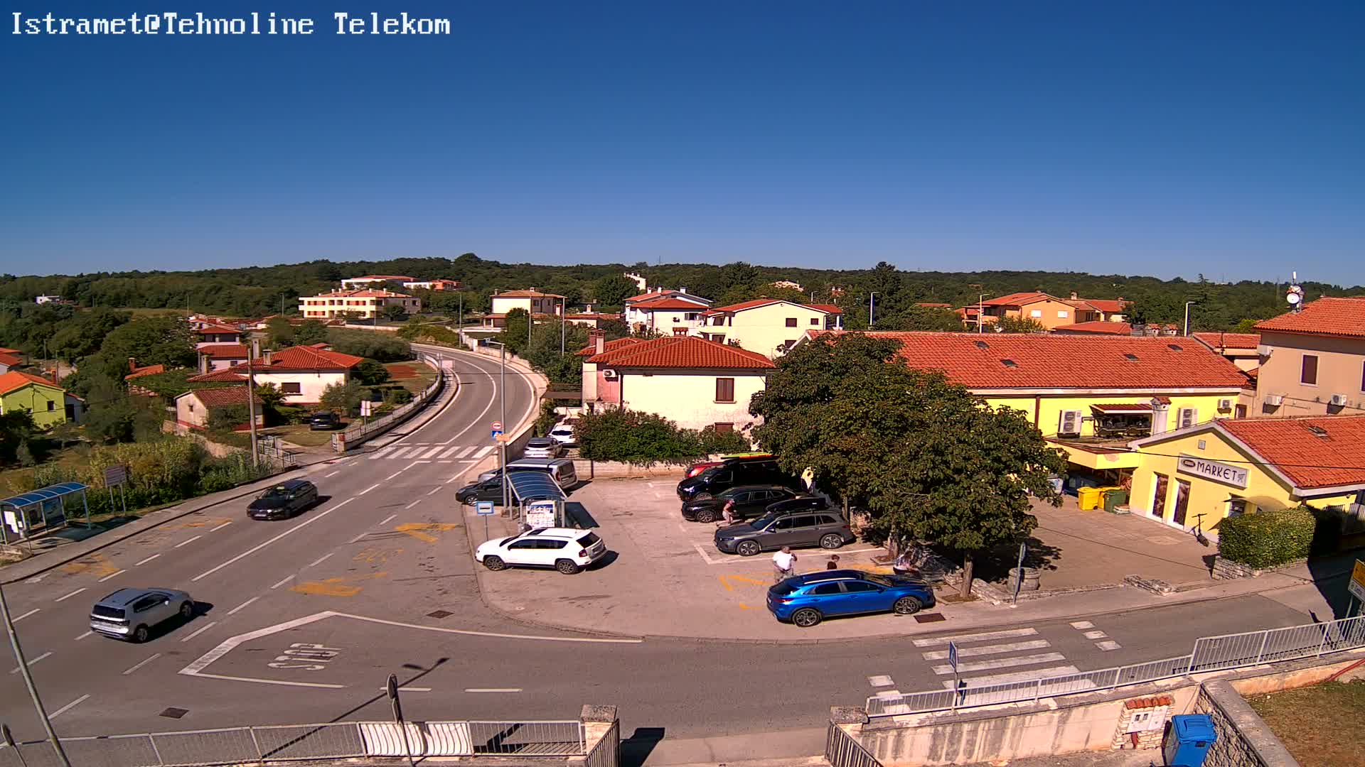 A sunny day reveals a small town with low-rise buildings, a road curving through it, and several cars parked in a lot near a small market.