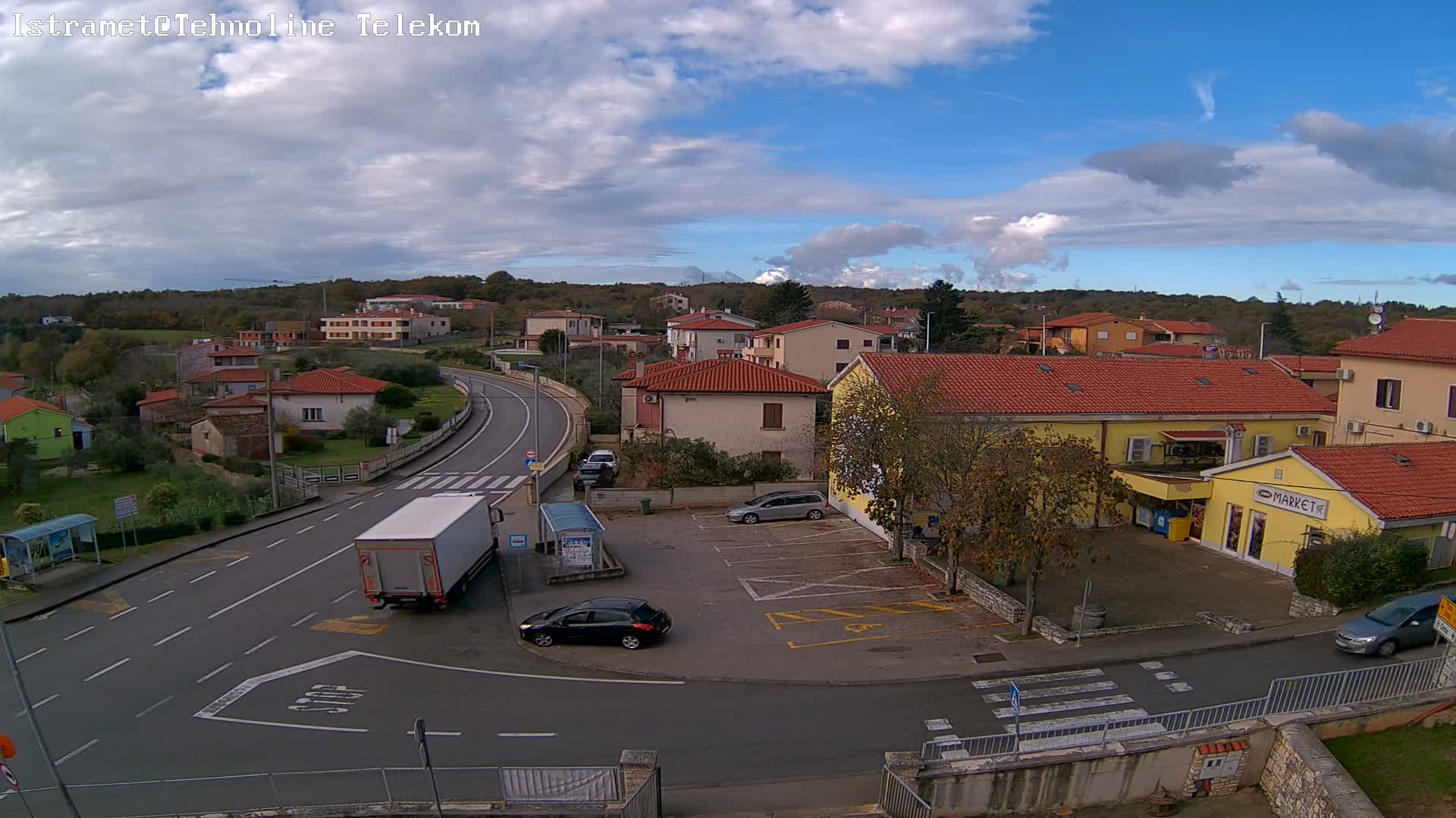 An elevated view captures a small town featuring numerous red-roofed houses, a winding road with vehicles, and a yellow market building nestled among trees, all under a bright, partly cloudy sky.