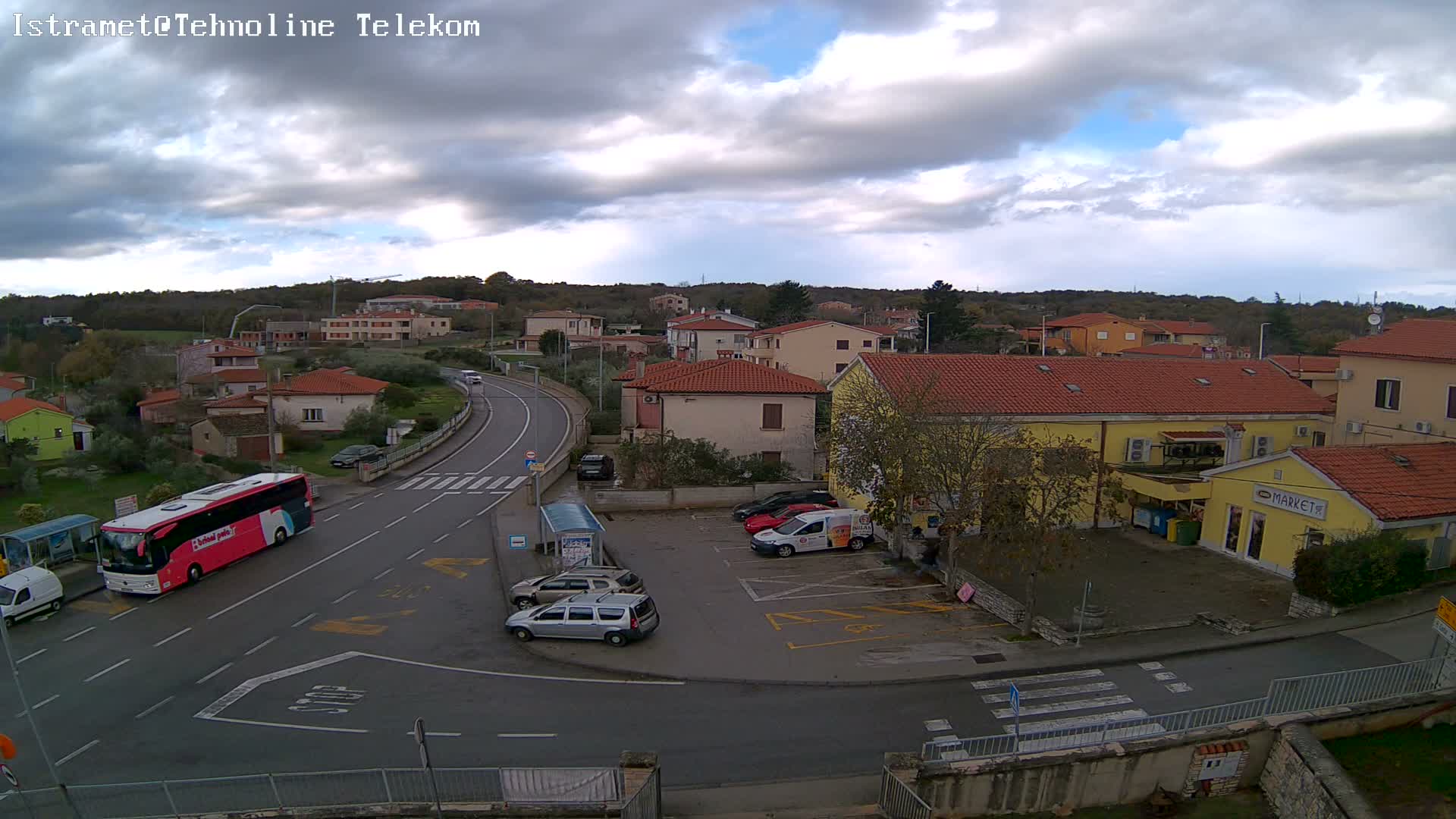An elevated view captures a small town featuring numerous red-roofed houses, a winding road with vehicles, and a yellow market building nestled among trees, all under a bright, partly cloudy sky.
