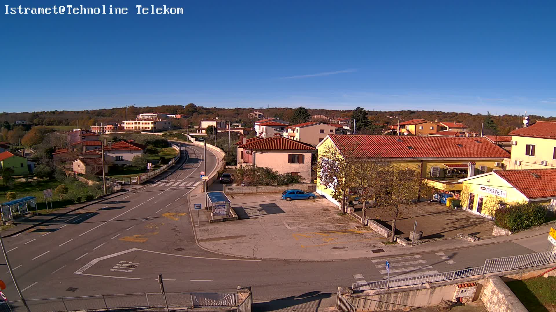 A sunny, clear day over a quiet residential area features a winding road with a bus stop and a parking lot, surrounded by houses with red tile roofs and some trees, with a market visible on the right and a wooded hill in the background.