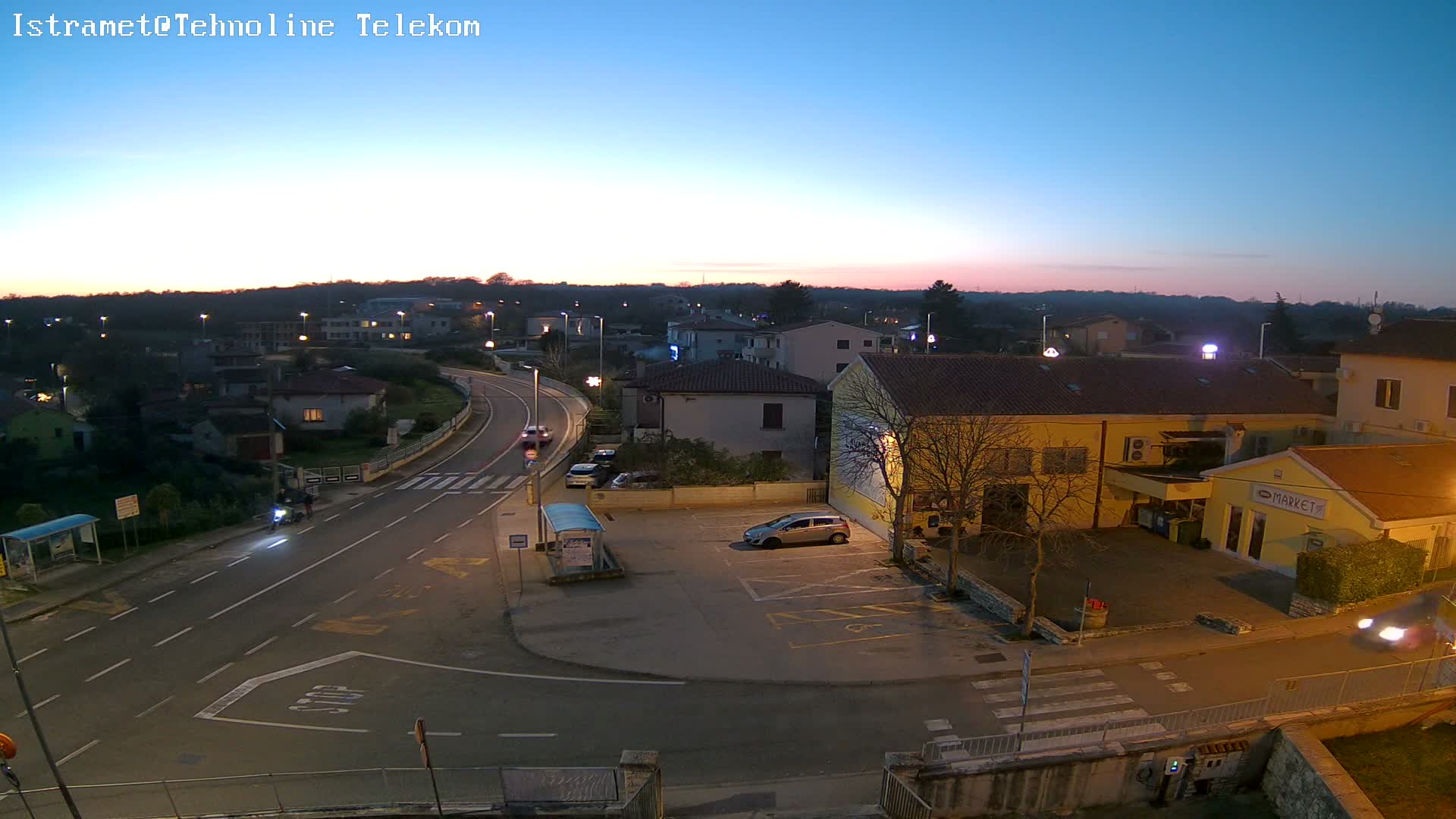 A sunny, clear day over a quiet residential area features a winding road with a bus stop and a parking lot, surrounded by houses with red tile roofs and some trees, with a market visible on the right and a wooded hill in the background.