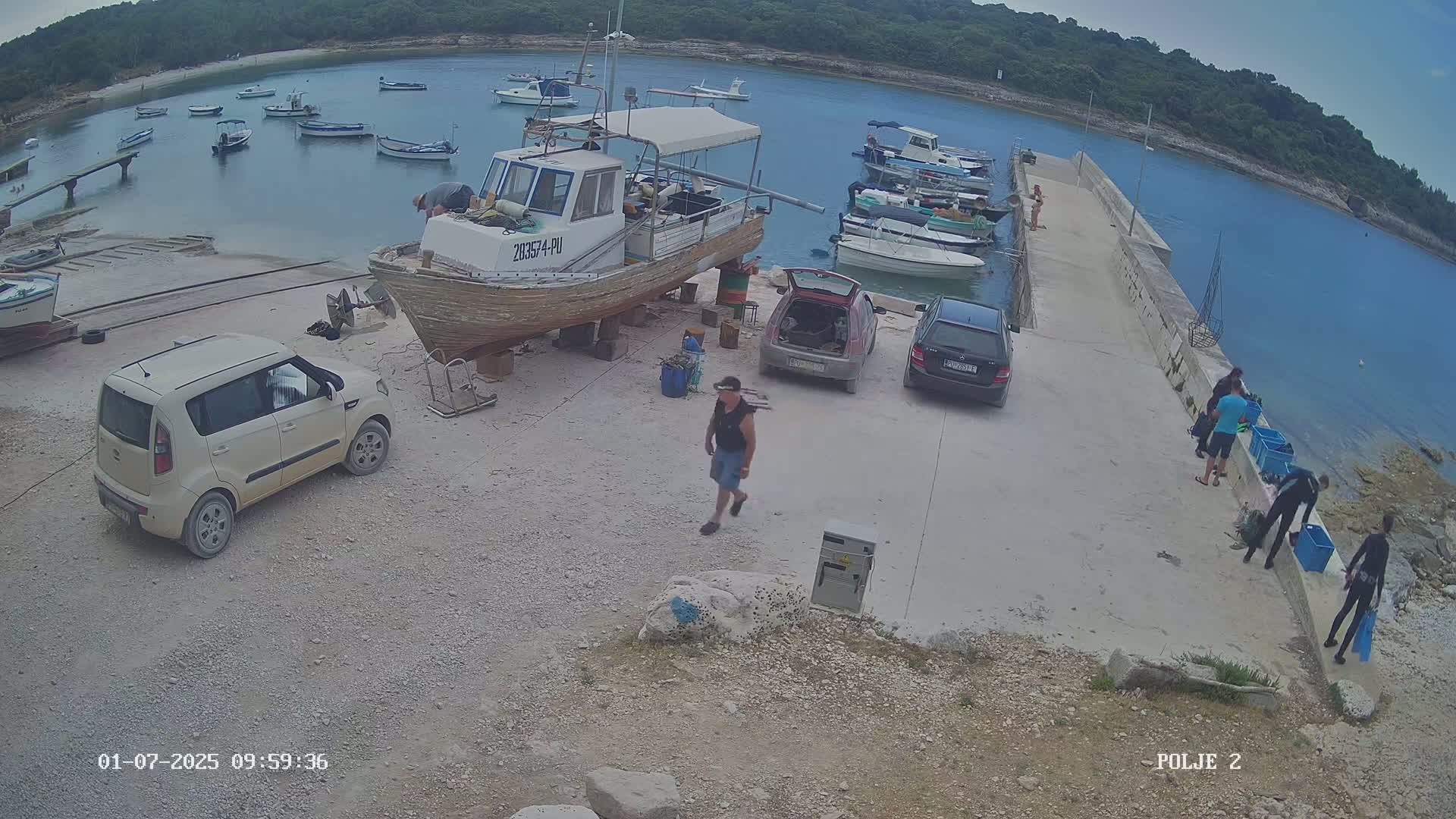 A small harbor with numerous boats, some cars parked nearby, and several people on a pier under a partly cloudy sky.