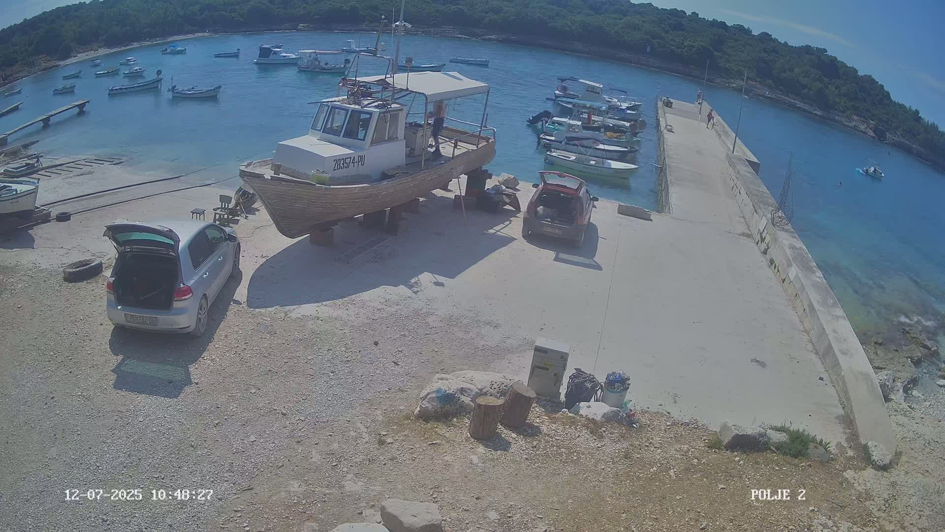A small harbor with numerous boats, some cars parked nearby, and several people on a pier under a partly cloudy sky.