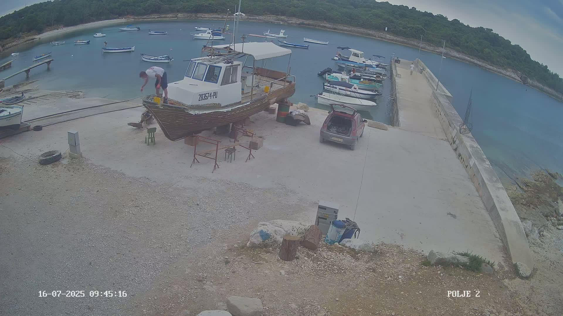 A man is working on a boat on a rocky shore next to a calm bay with numerous other boats, under an overcast sky.