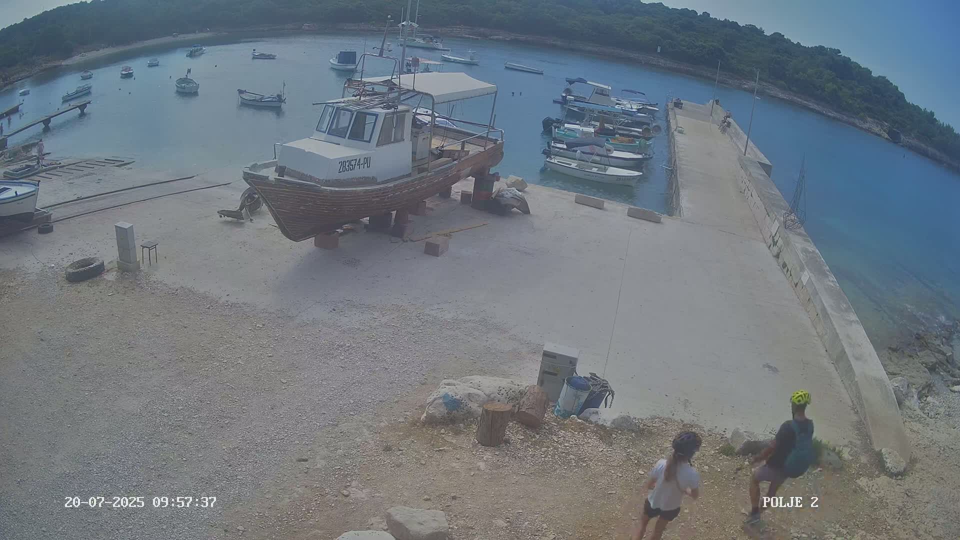 A small harbor with numerous boats, including a larger one on land supported by blocks, is seen on a sunny day, with two people standing on a nearby rocky shore.