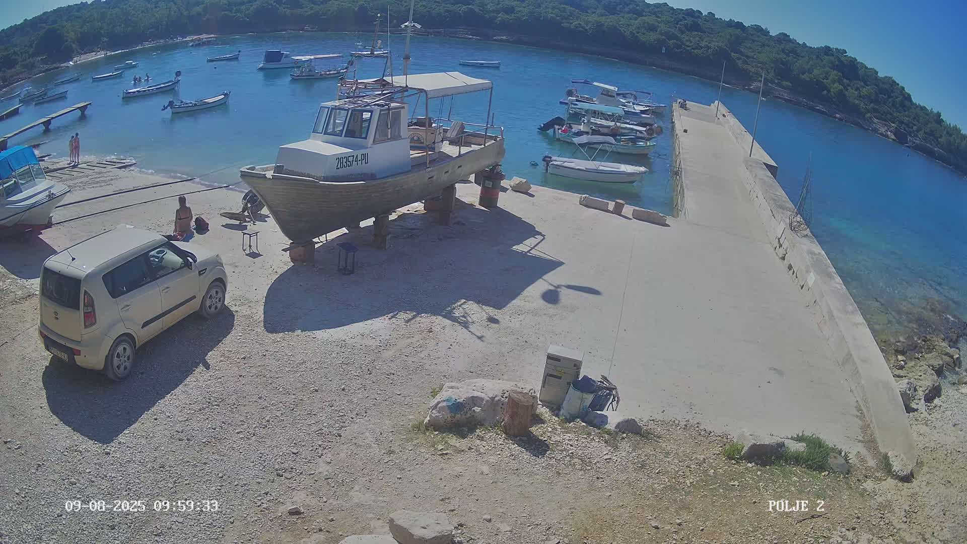 A beige car is parked near a boat on supports in a small harbor filled with other boats on a sunny day.