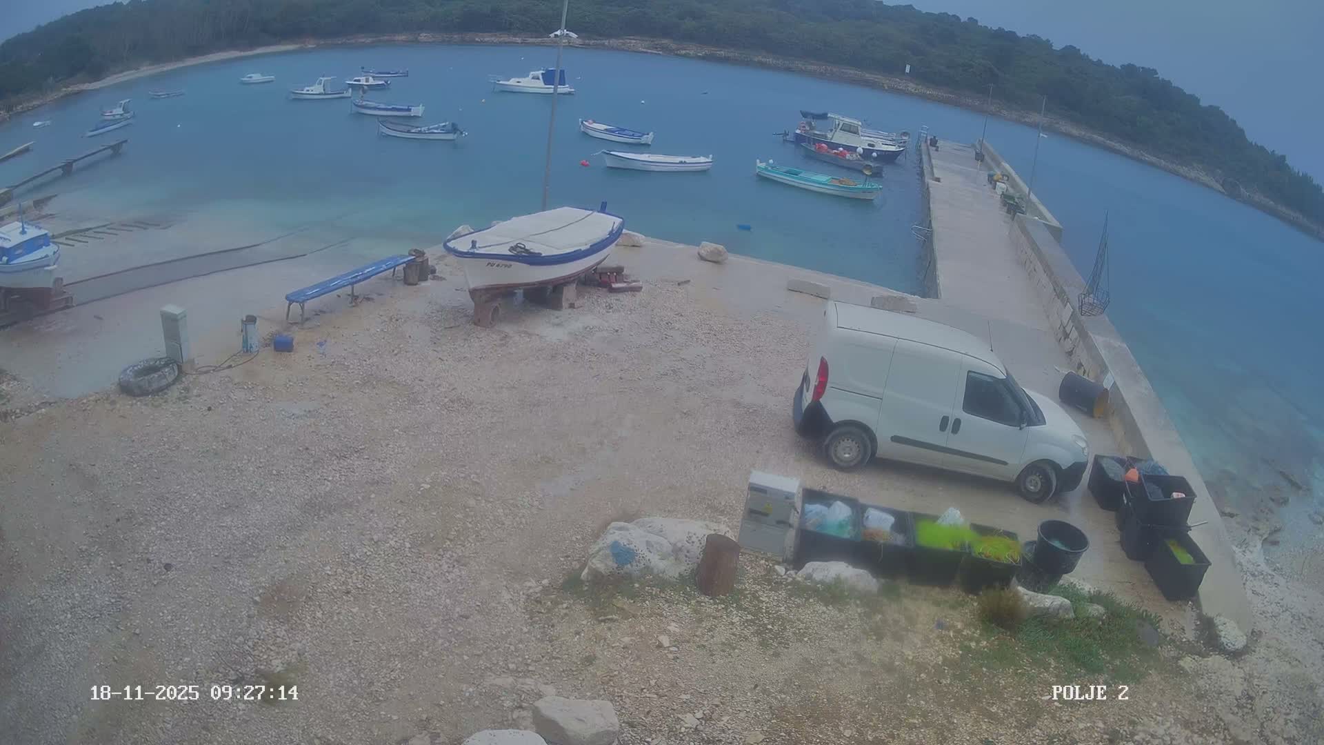 A small harbor with numerous boats afloat and two boats on the gravel shore, alongside a white van parked on a concrete pier, is visible under an overcast sky.
