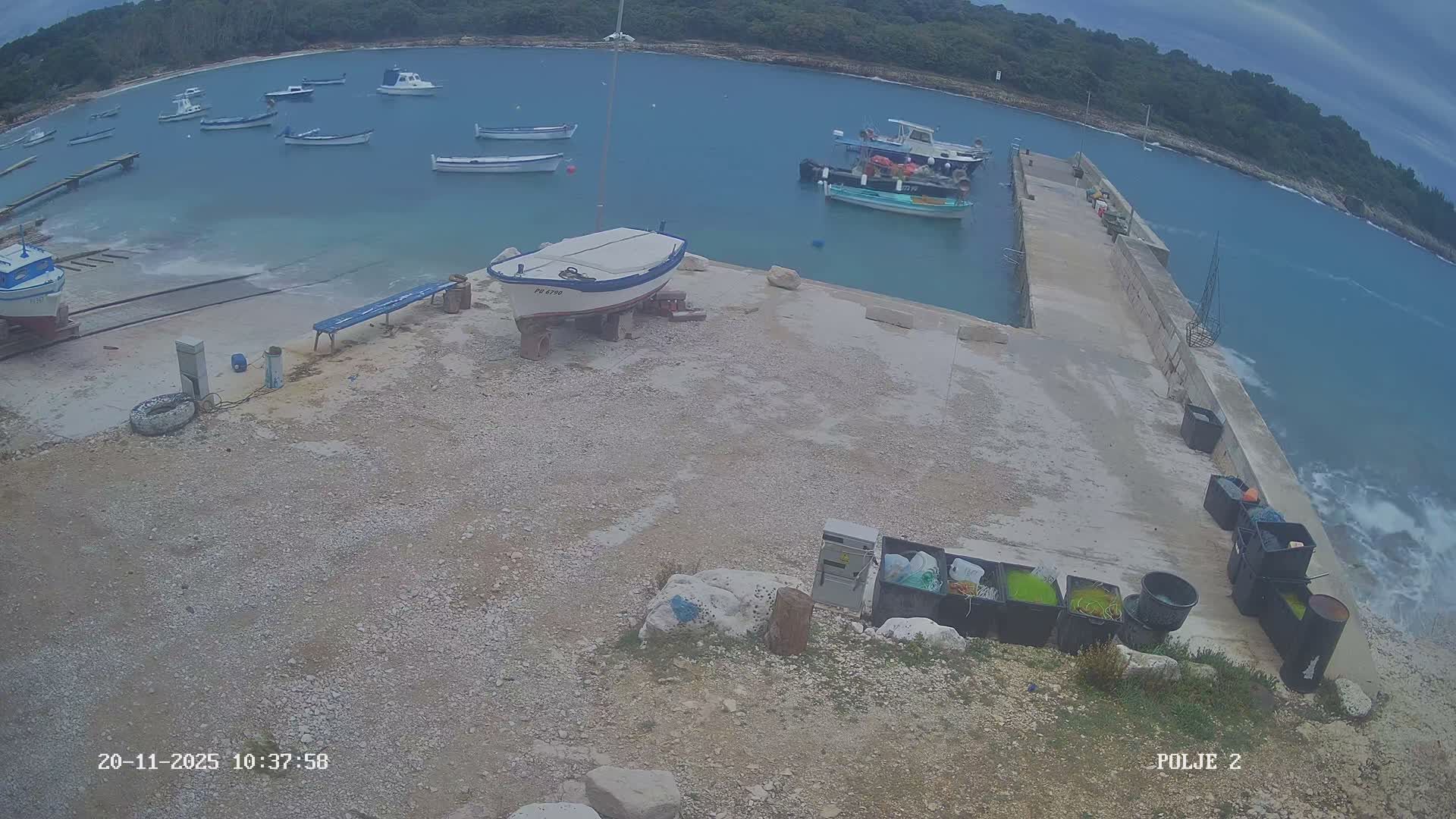 On an overcast day, a concrete pier extends into a bay with numerous boats, while two boats are dry-docked on the gravelly shore and choppy water churns near the outer pier.