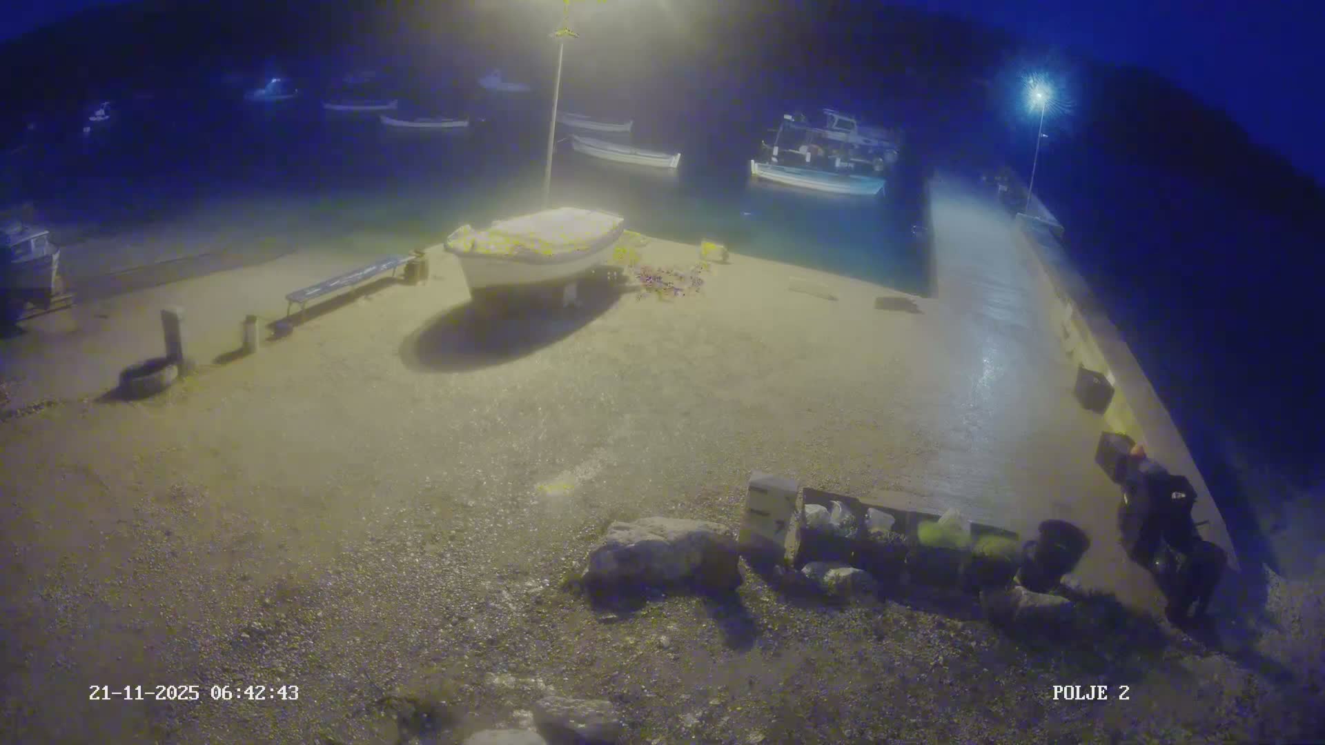 On an overcast day, a concrete pier extends into a bay with numerous boats, while two boats are dry-docked on the gravelly shore and choppy water churns near the outer pier.