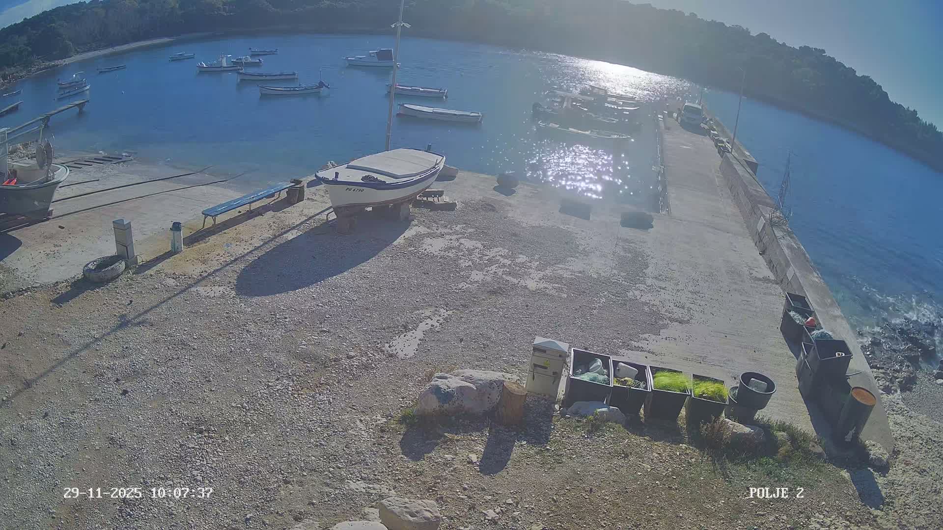 Under clear, sunny skies, a tranquil bay features numerous small boats moored in the sparkling water, alongside a gravel shore with a boat pulled ashore and a concrete pier extending into the water with fishing gear stored nearby.