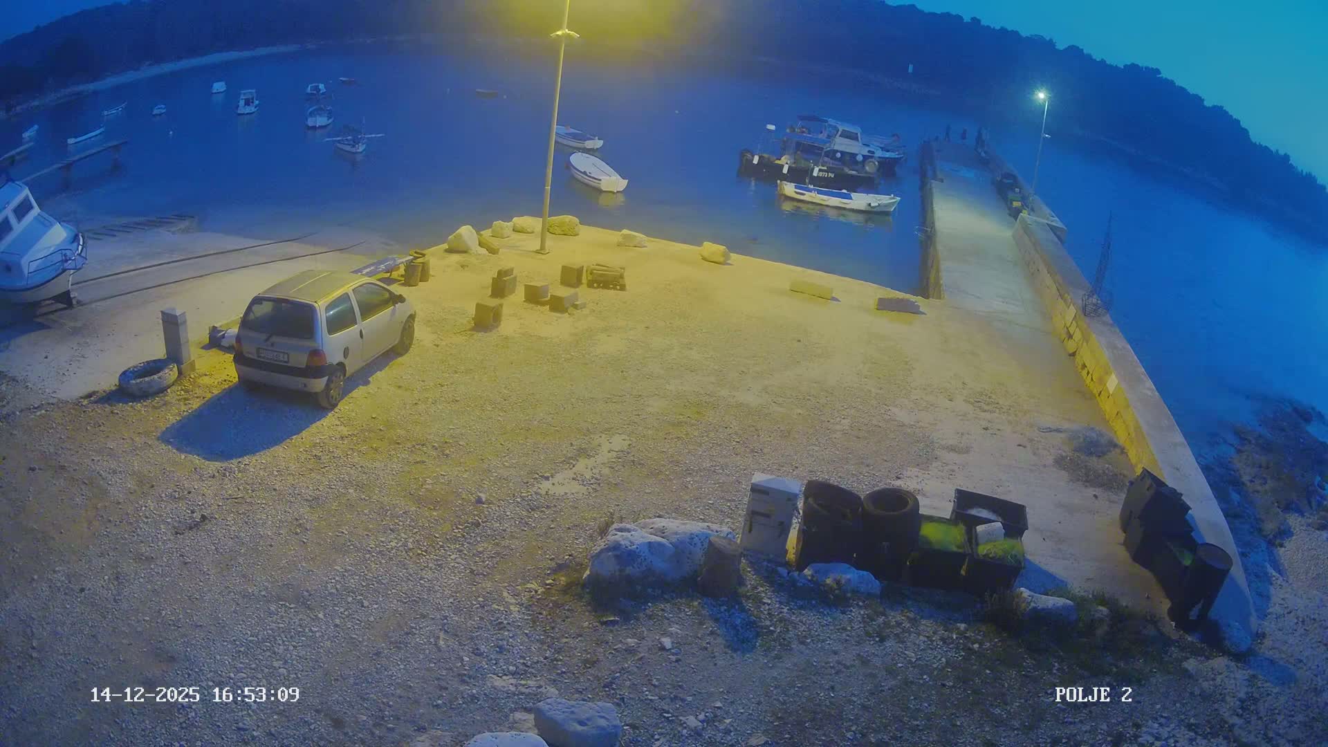 Under clear, sunny skies, a tranquil bay features numerous small boats moored in the sparkling water, alongside a gravel shore with a boat pulled ashore and a concrete pier extending into the water with fishing gear stored nearby.