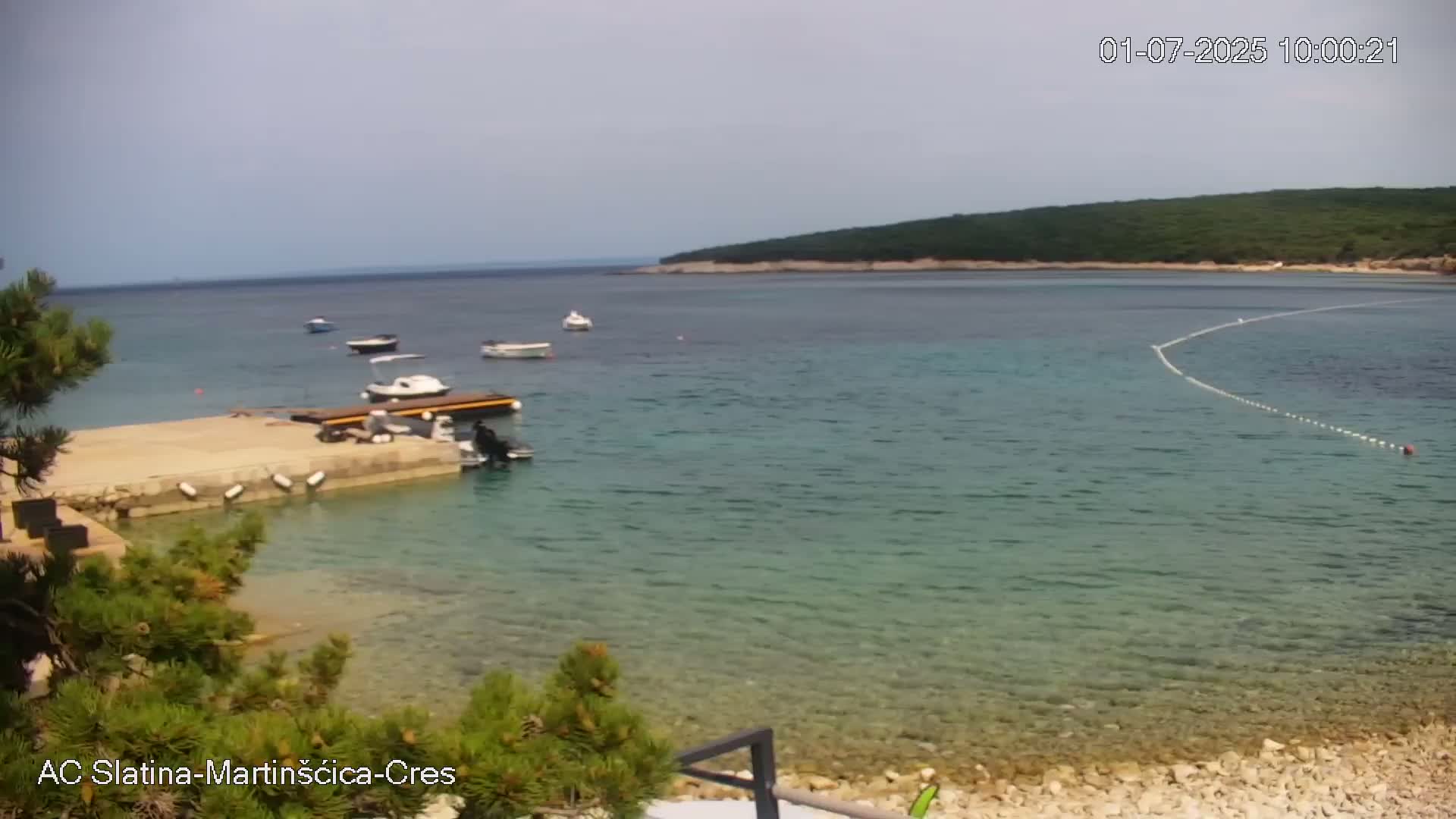 A calm, clear blue sea laps a pebbled shore near a small dock with several boats moored, backed by green hills under a mostly clear sky.