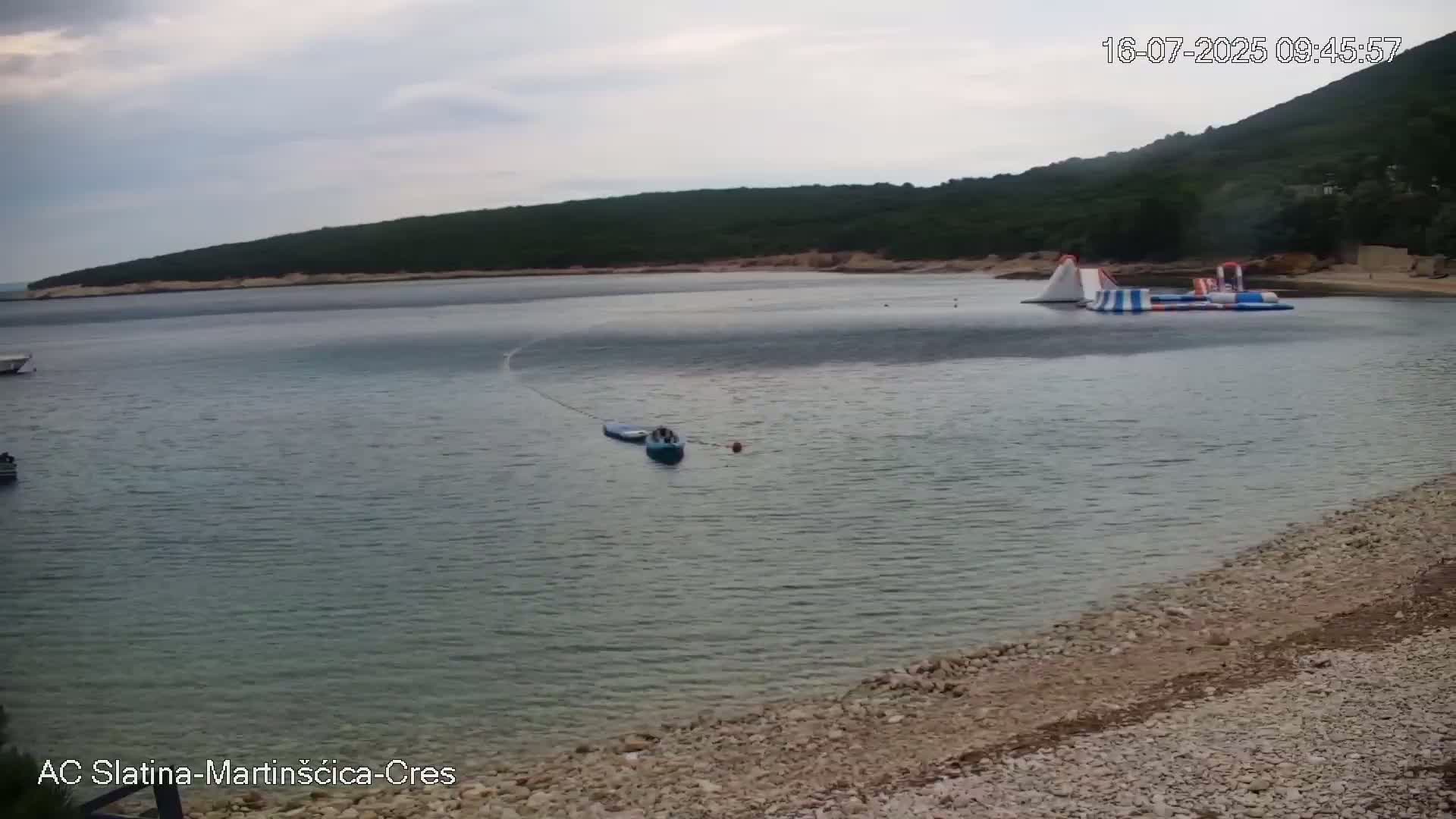 A calm, overcast day at a bay with a rocky shore shows a small inflatable water park and a few boats.