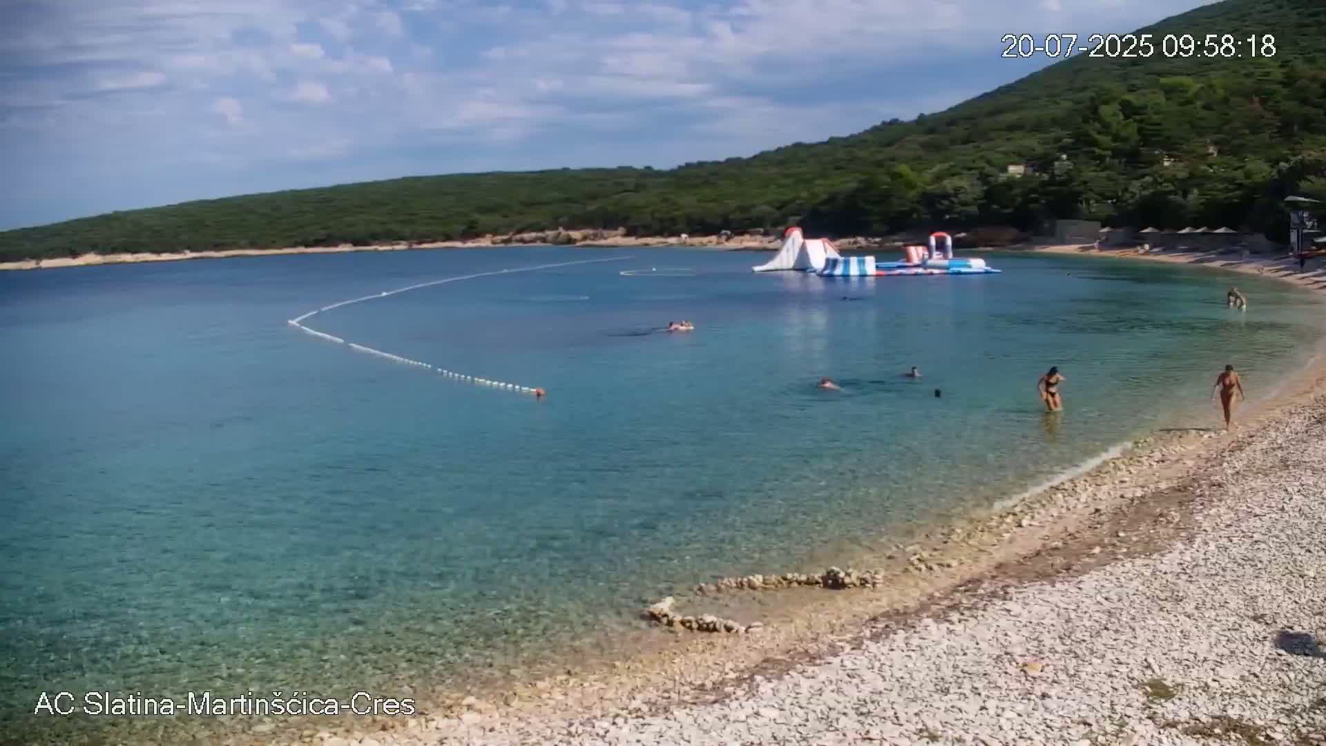 A sunny day at a pebble beach with clear, shallow turquoise water, featuring an inflatable water park and several people swimming and sunbathing.