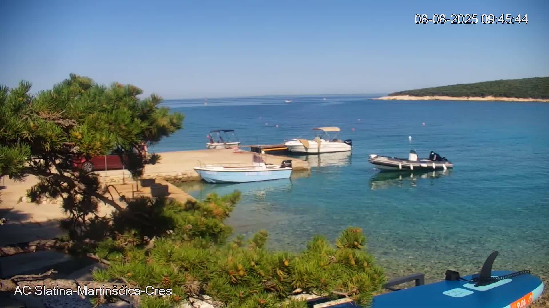 Several small motorboats are moored at a calm, clear, blue-water shoreline on a sunny day, near a small island visible in the distance.