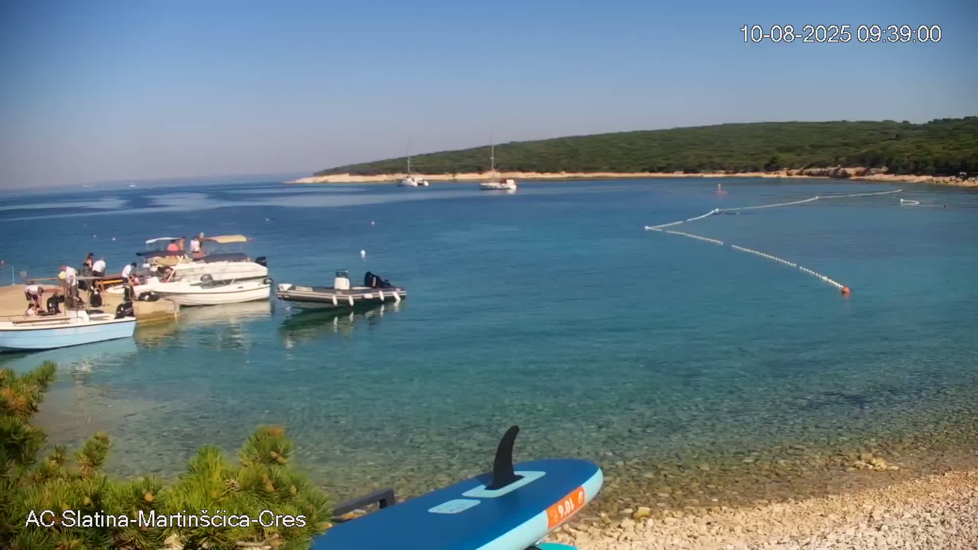 Several boats are moored near a rocky shore on a clear, sunny day, with a calm, turquoise sea and a green, hilly island in the background.
